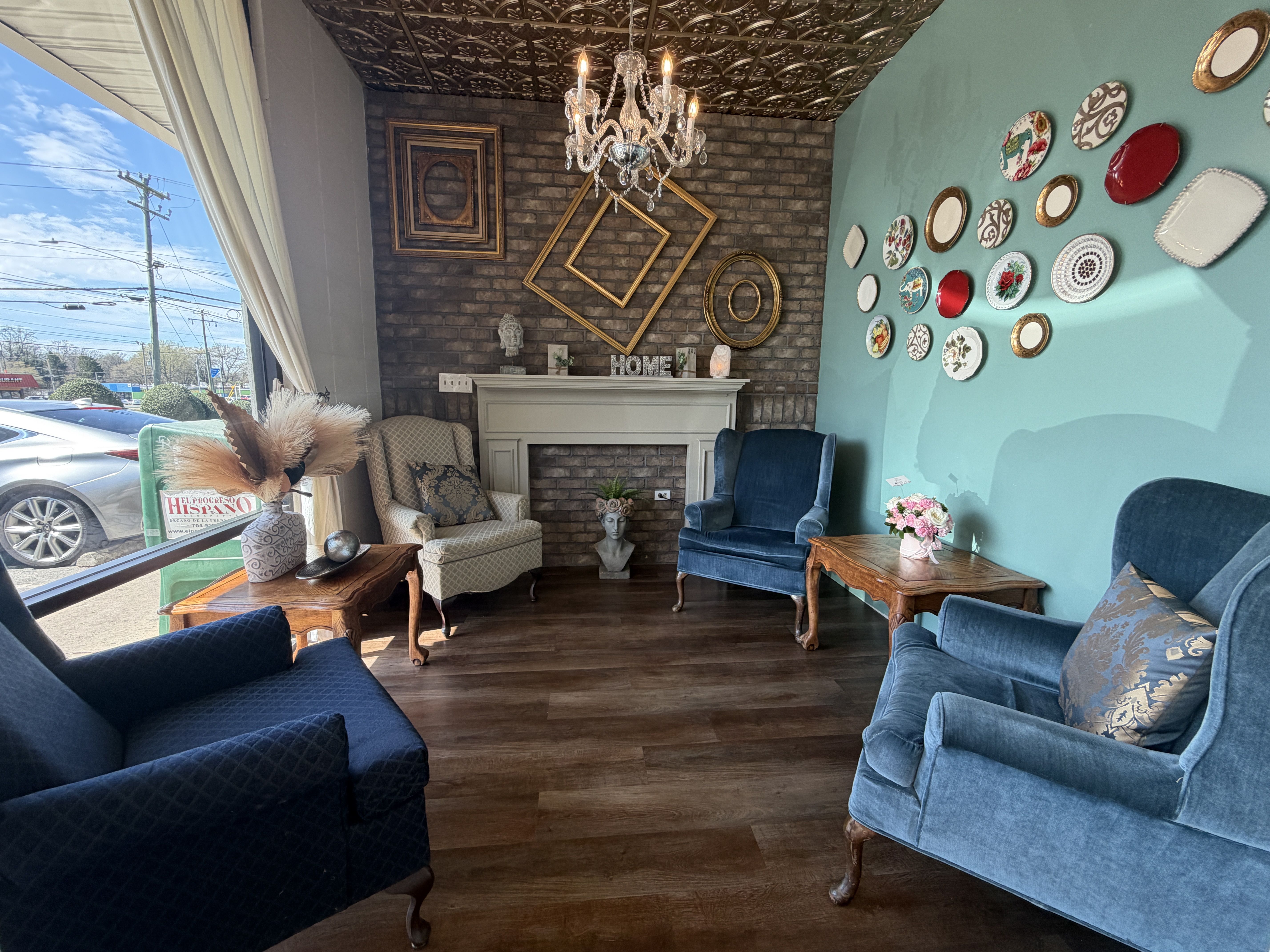 Cozy showroom living room with a teal wall decorated by plates, a white mantel over a brick fireplace, a crystal chandelier, blue velvet chairs, wooden coffee tables, and a large window with curtains.