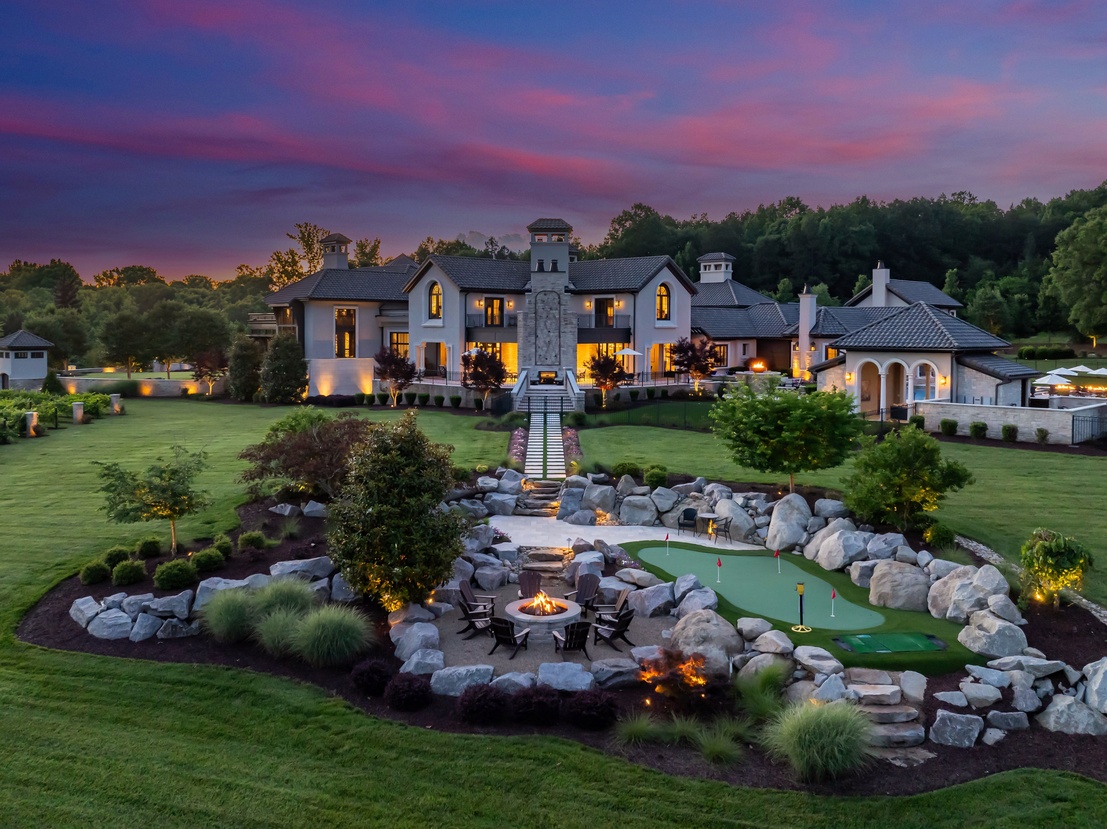 Large luxurious house with lit windows at dusk, surrounded by green lawn, garden with rocks, trees, fire pit seating area, and small putting green under a purple and blue sky.