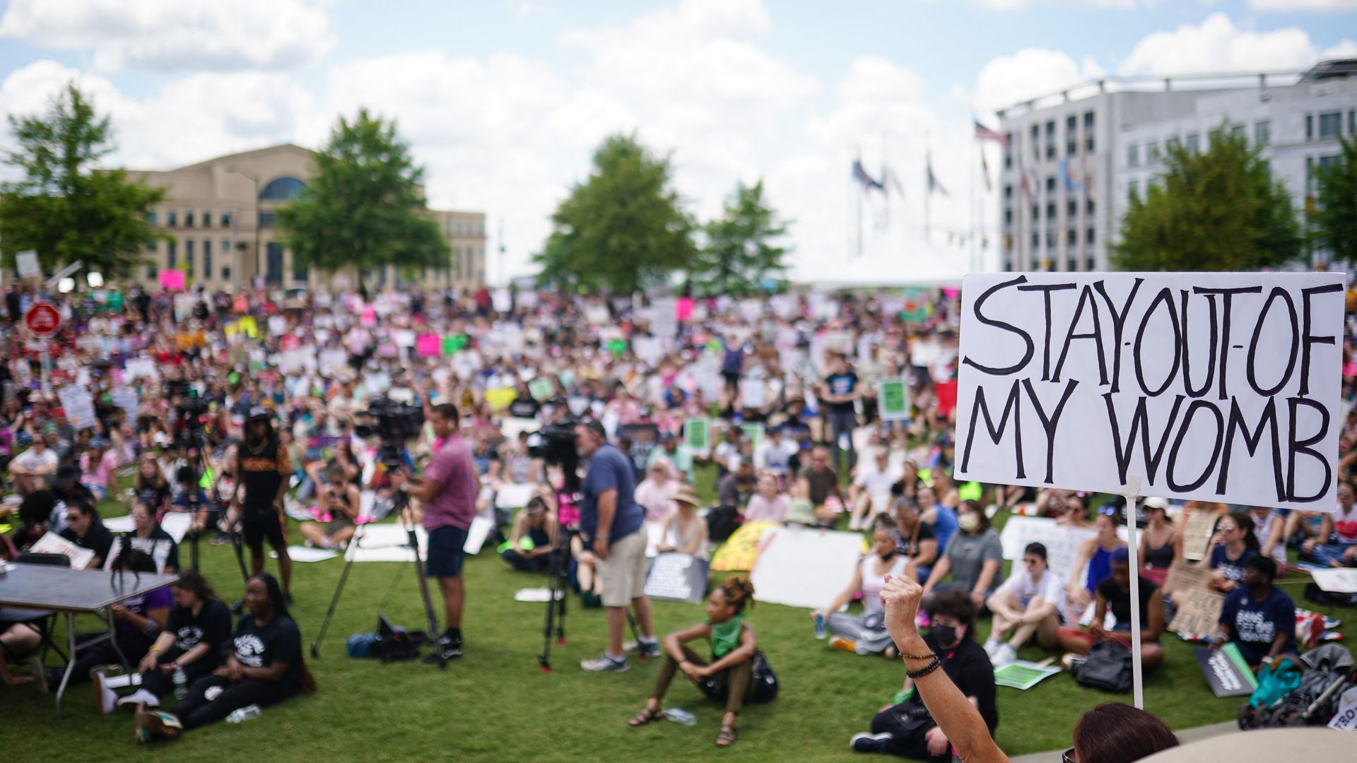 Photo of a sign that says "Stay out of my womb" against the backdrop of a crowd of protesters sitting on a green lawn