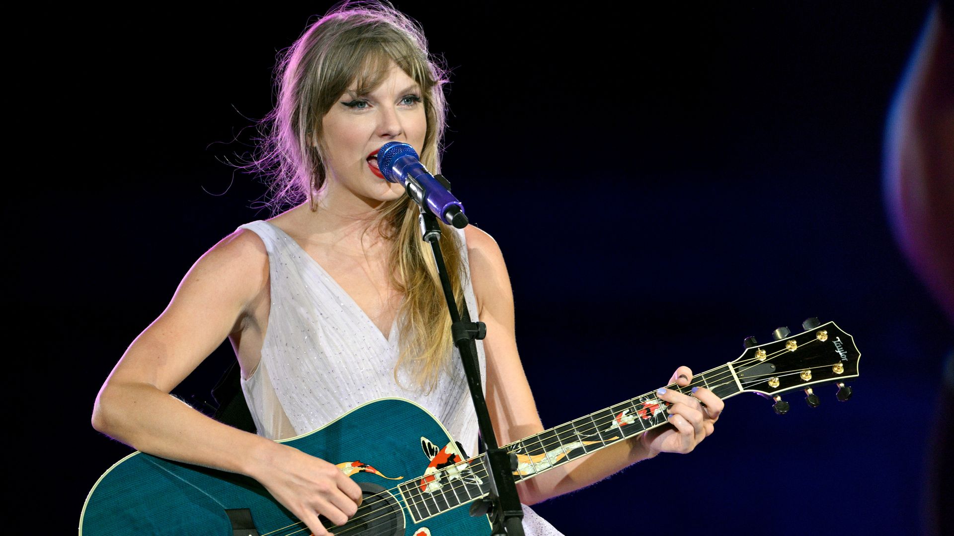 Singer Taylor Swift wears a sleeveless white dress while playing a blue guitar and singing. Her blond-ish hair hangs over her left shoulder and lights create a slight purple outline around her head.