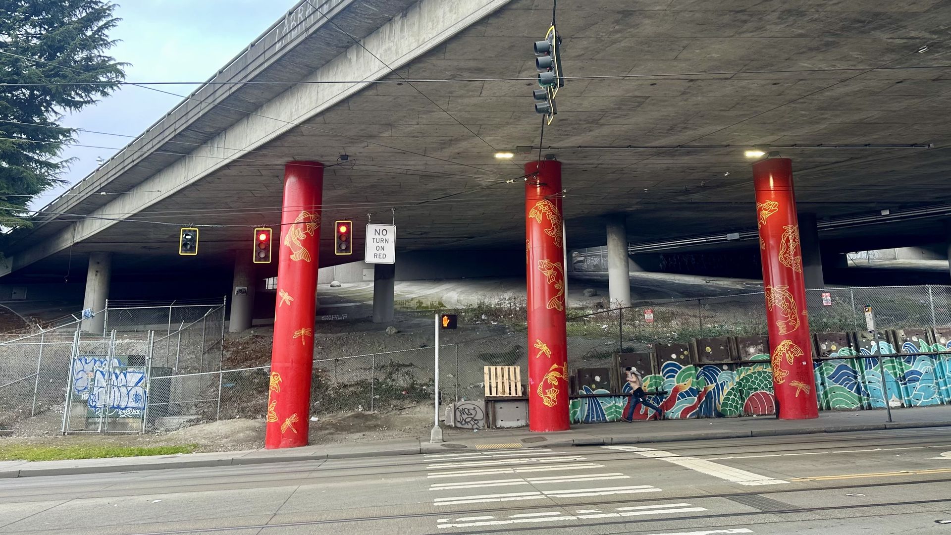 Three red and yellow colored support columns hold up a freeway overpass, with chain link fencing surrounding some areas under the freeway and a surface-level road in the foreground.