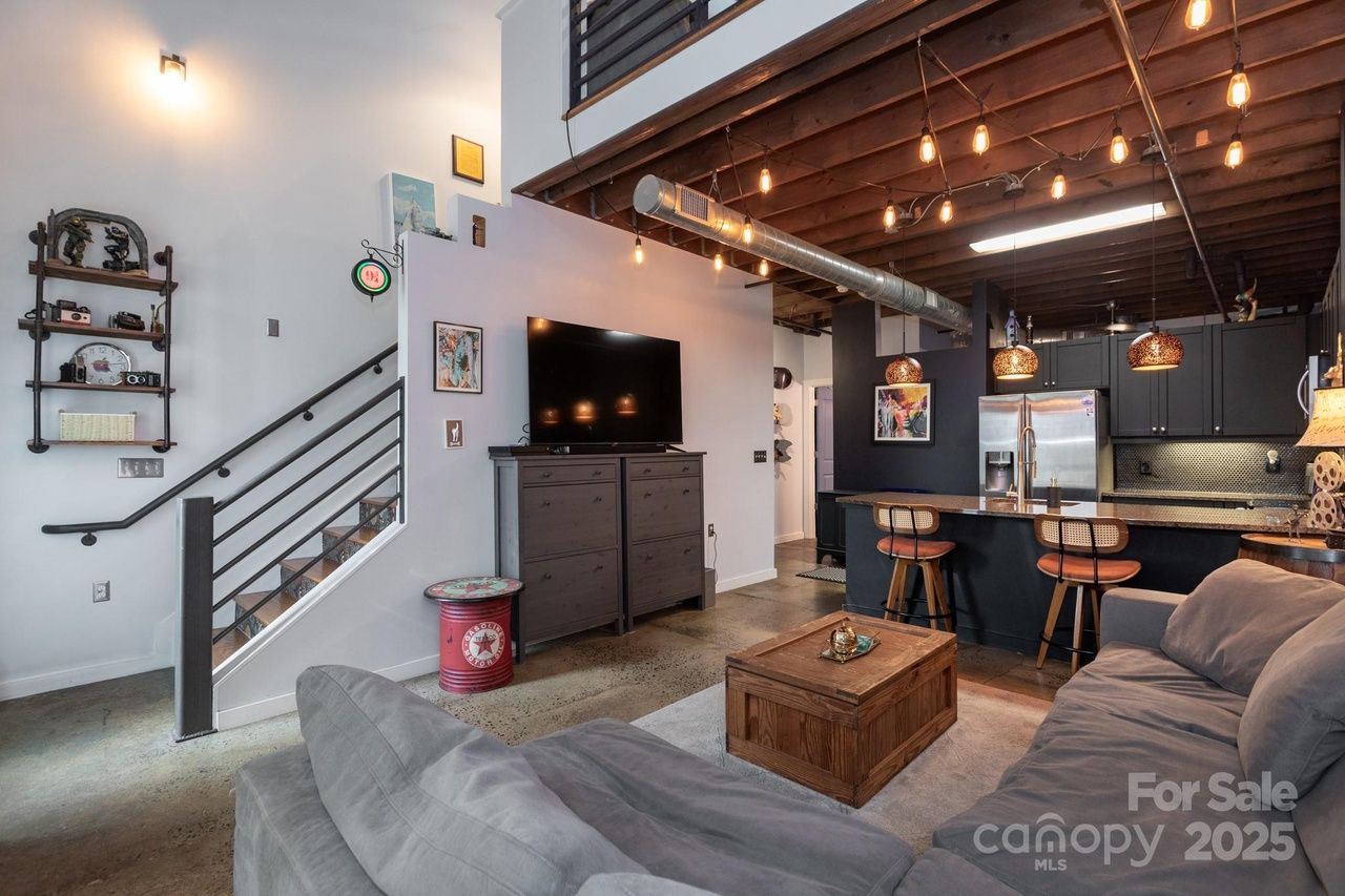 Modern living room with gray couch, wooden coffee table, black TV stand, and open kitchen with black cabinets, stainless steel appliances, exposed wooden ceiling beams, and hanging lights.