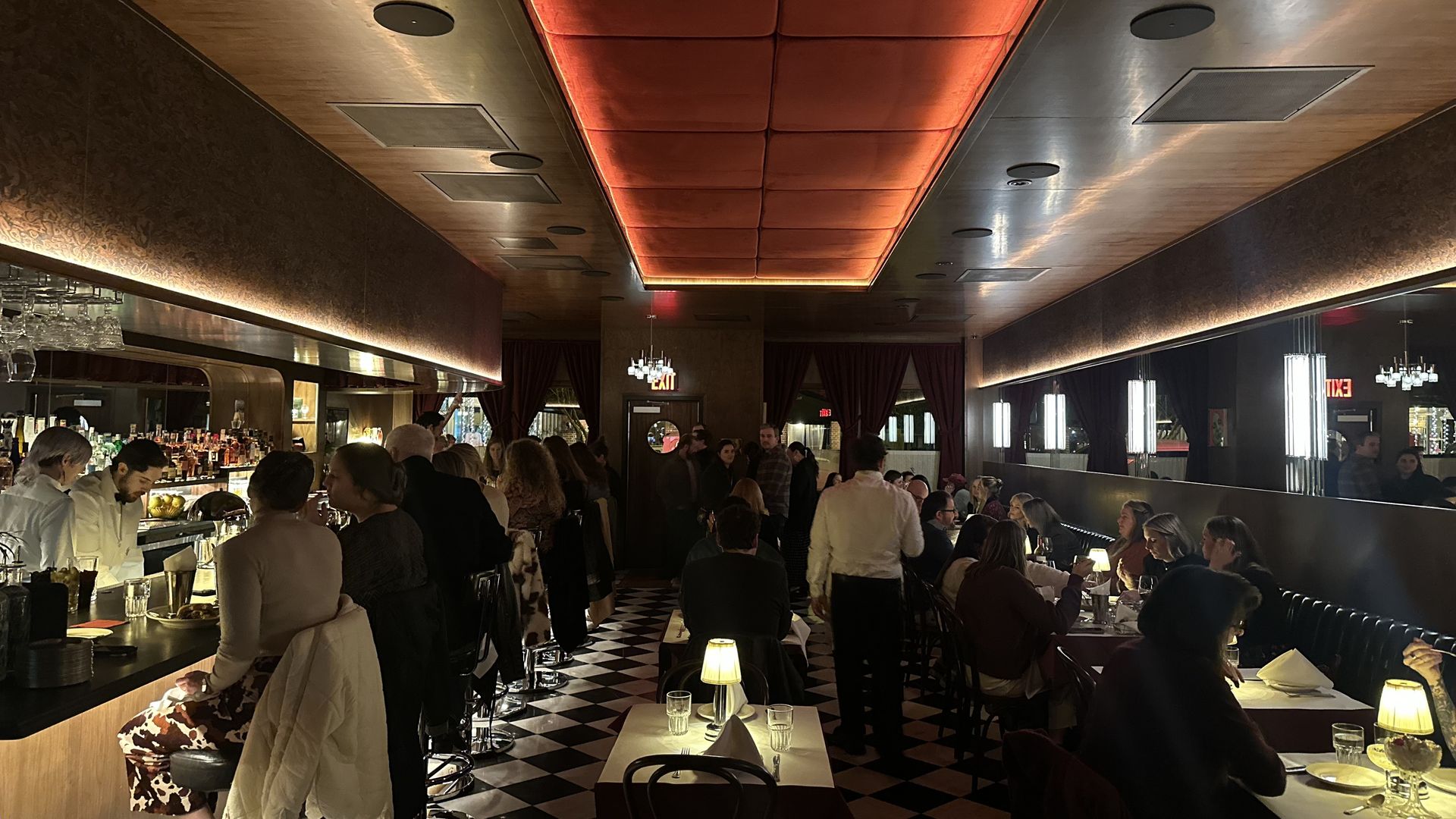Dimly lit restaurant interior with black and white checkered floor, red ceiling panel, long bar on left with seated patrons, tables with lamps on right, and groups dining.