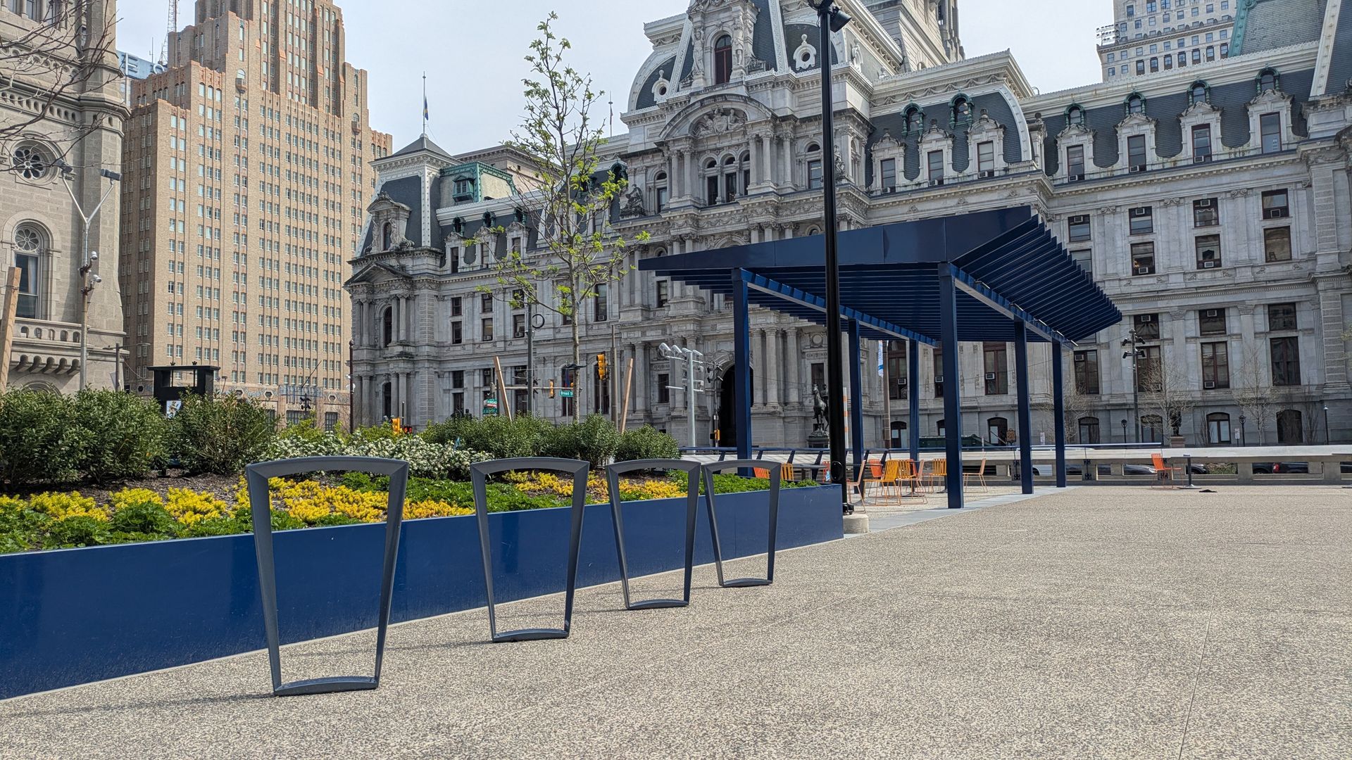 Urban plaza with an ornate historic building in the background, a blue pergola with orange chairs, a long blue planter bed with yellow flowers, and dark curved metal frames in the foreground.