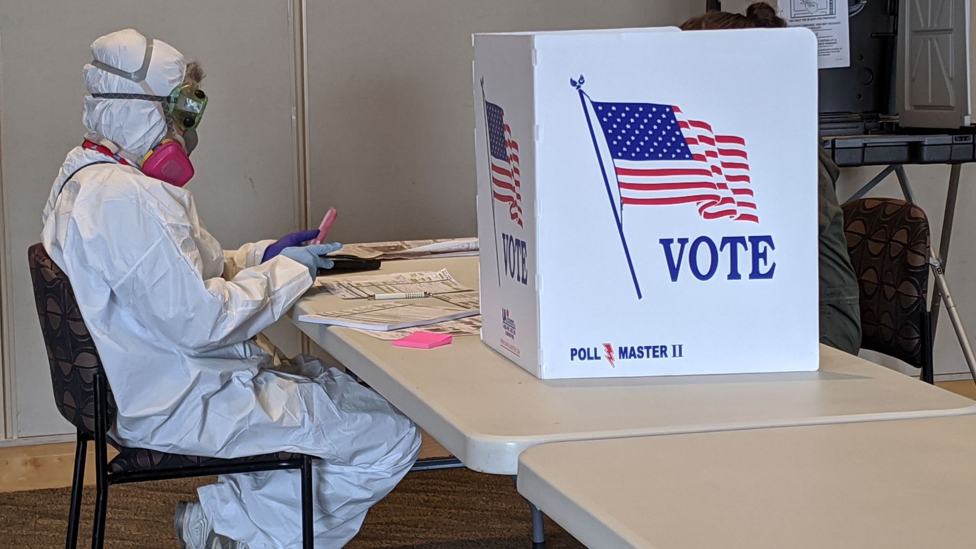 In this image, a person in a hazmat suit and gas mask sits in front of a voting booth