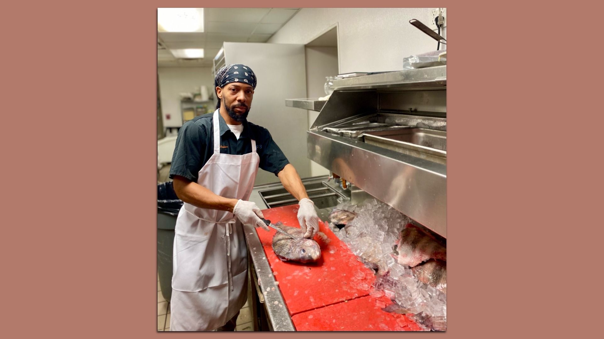 Man in white apron and bandana fillets a fish on a red cutting board next to a counter with ice and more fish in a commercial kitchen.