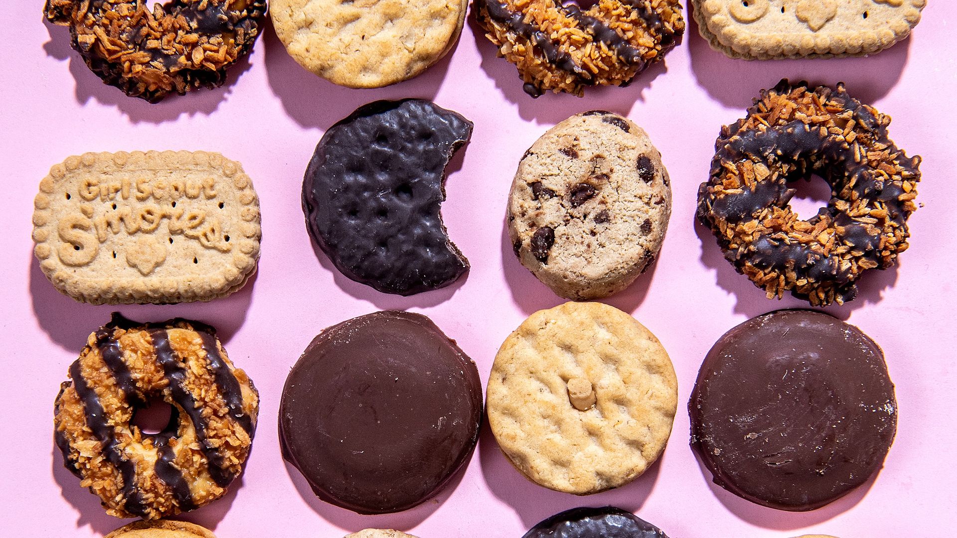 Girl scout cookies laid out in rows on a pink background.