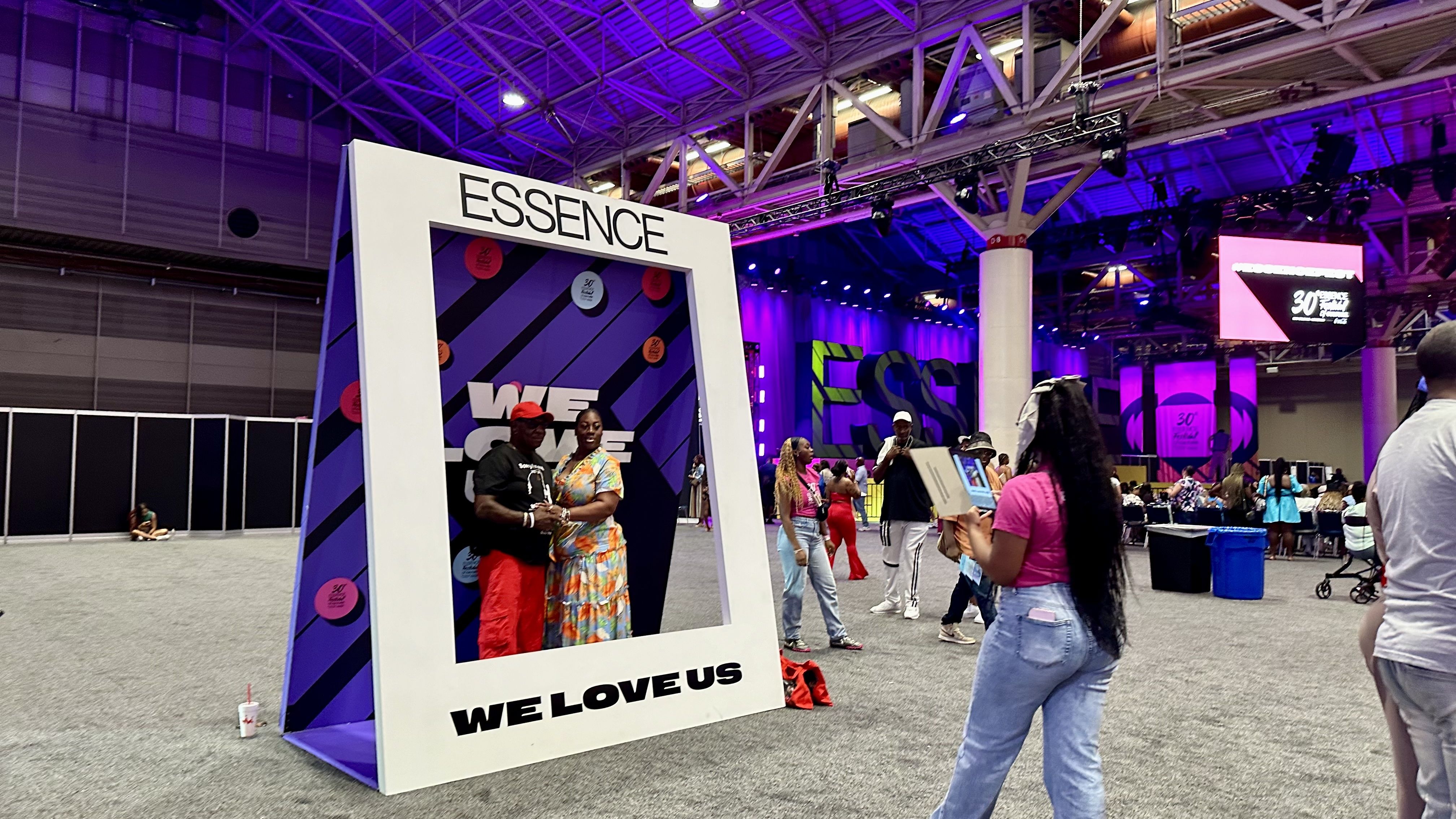 Indoor convention hall with purple lighting. A large white frame says "ESSENCE" at the top and "WE LOVE US" at the bottom. Two people pose inside; other attendees and vibrant displays in the background.