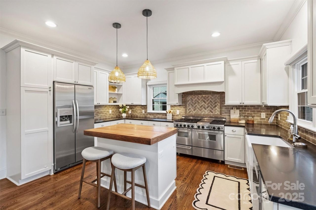 Bright modern kitchen with white cabinets, stainless steel appliances, wood island with two gray stools, wooden floor, beige herringbone tile backsplash, and two woven pendant lights.