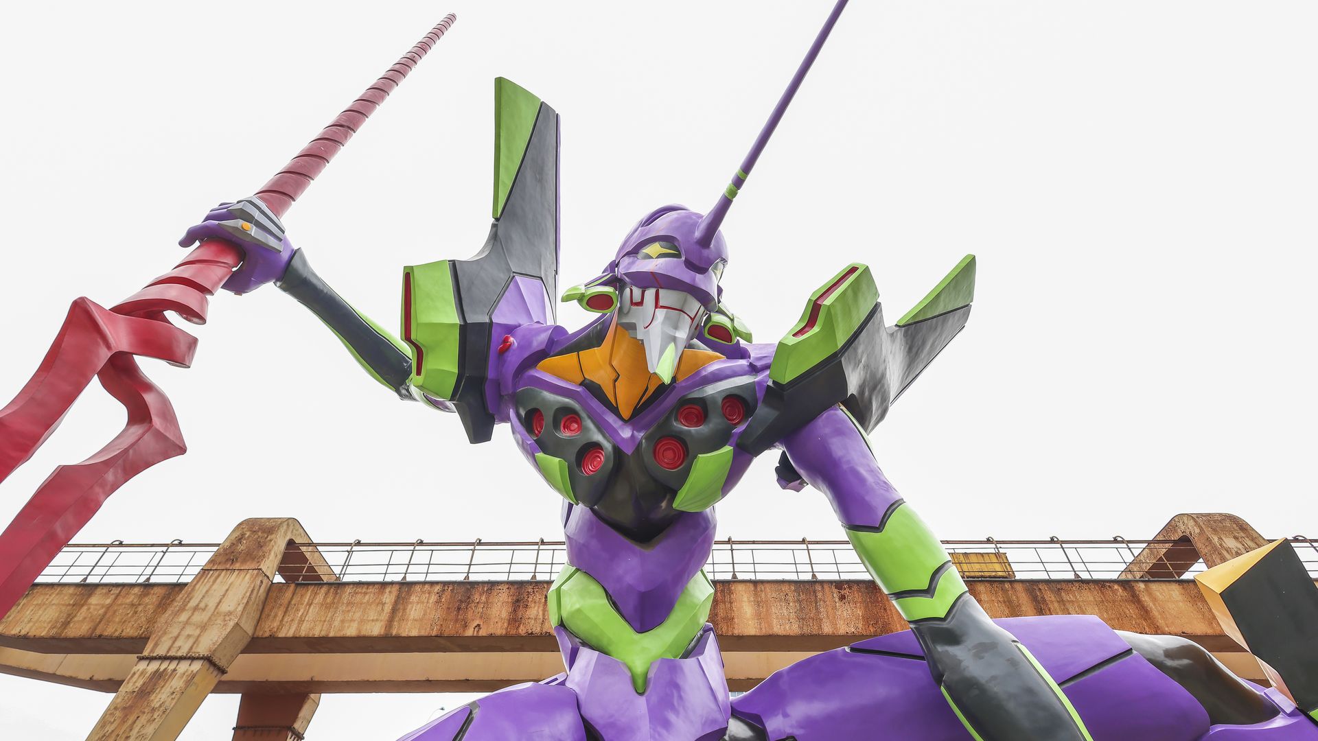 A life-size sculpture of Evangelion Unit-01 in purple and green, wielding a long pink spear, standing on a rusted industrial bridge with a pale sky behind.
