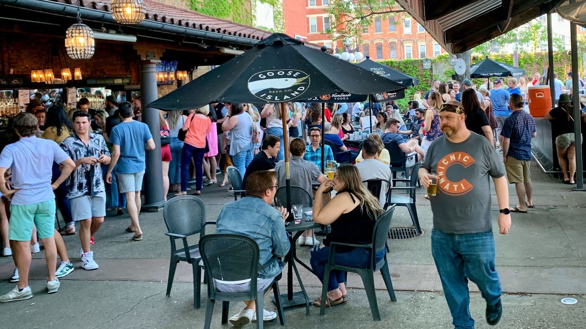 Bustling outdoor beer garden under a striped canopy with hanging lanterns and leafy trees. People mingle around tables beneath Goose Island umbrellas, brick buildings visible in the background.