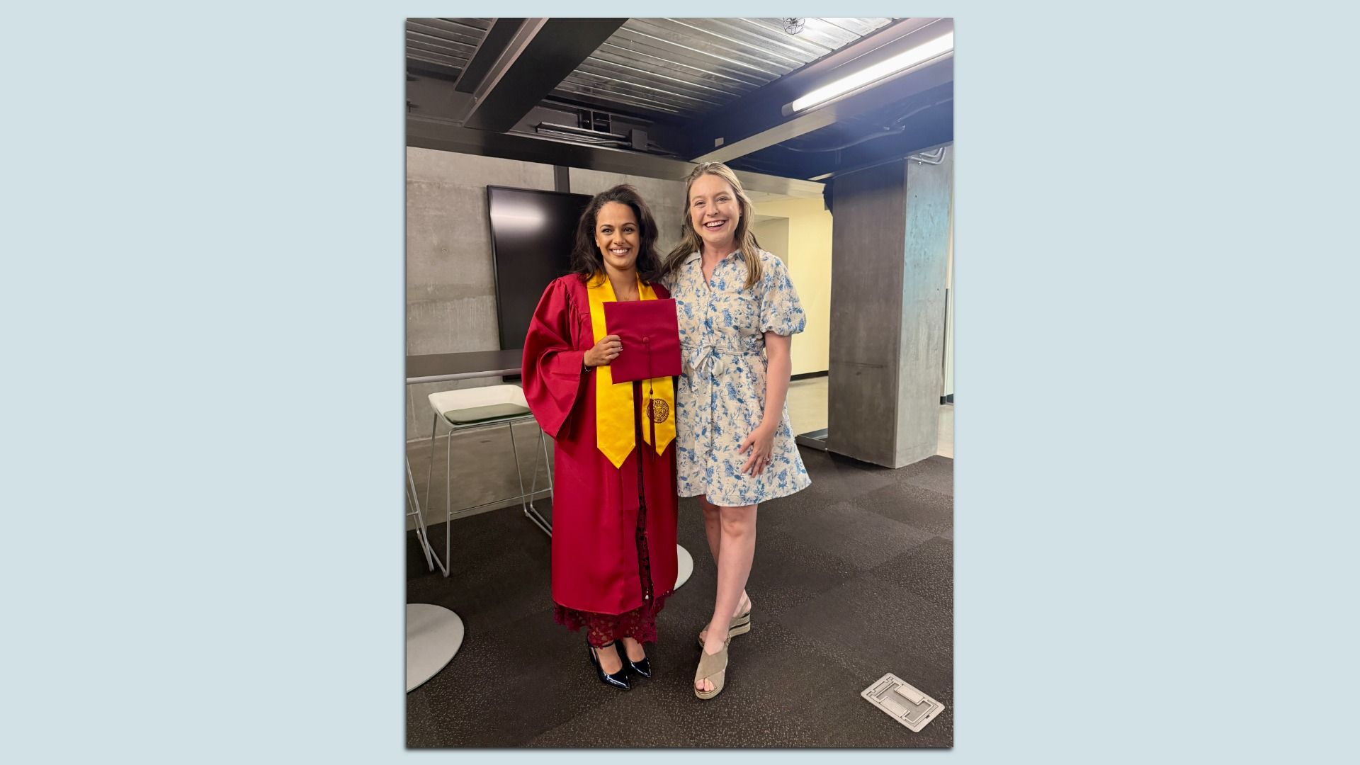 Two women pose in a gray, industrial hallway. One wears a red graduation gown with a yellow stole and holds a red diploma cover; the other is in a blue floral dress. Both are smiling.