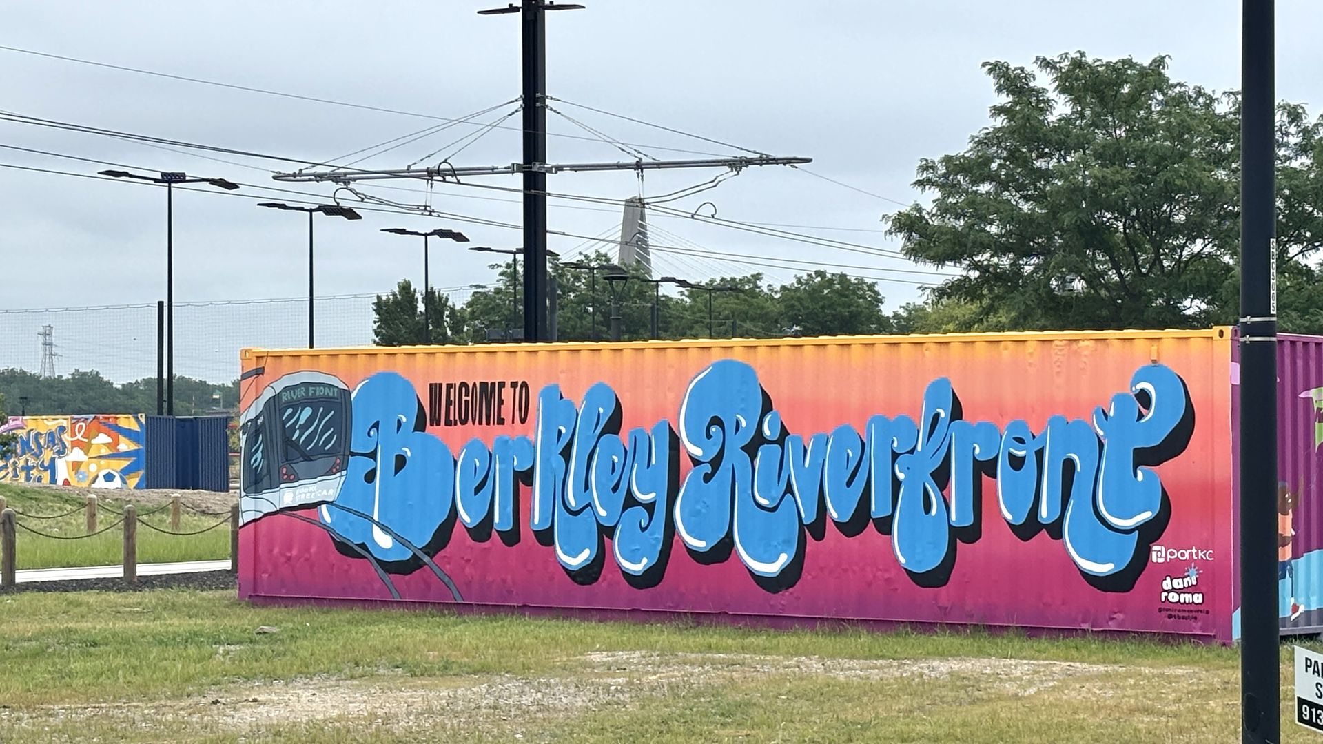 Bright orange graffiti-covered shipping container with large blue script text reading "Welcome to" and ending with "Riverfront"; yellow rim at top, a purple container to the right, and power lines with trees in the background.