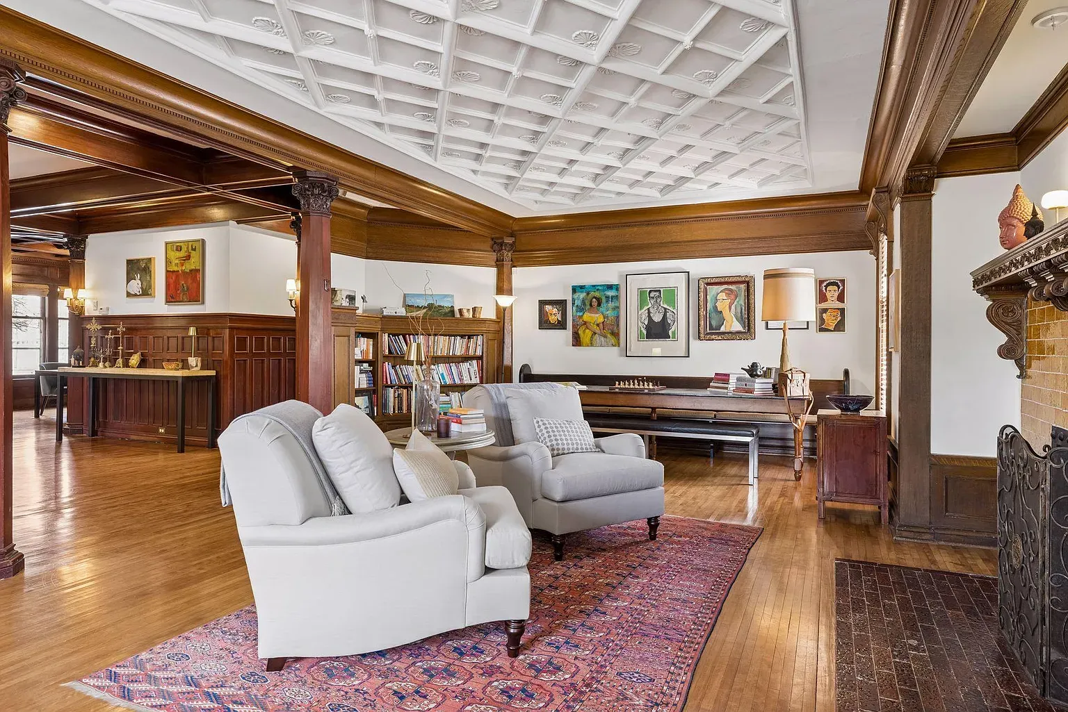 Spacious living room with white coffered ceiling, dark wood beams and columns, built-in bookshelves, and artwork. Two light gray armchairs rest on a red patterned rug beside a brick fireplace.