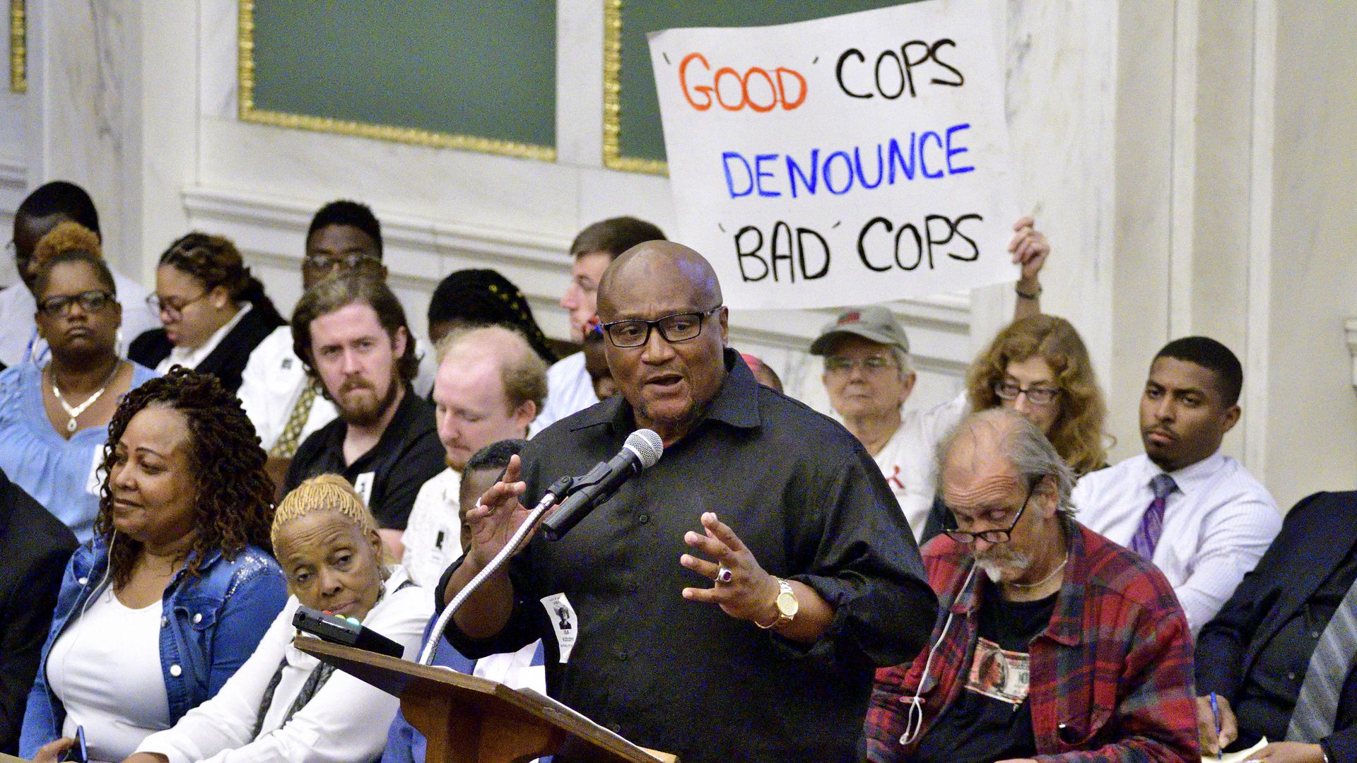 In this image, a man speaks at a podium while a woman holds a sign behind him saying "Good cops denounce bad cops""