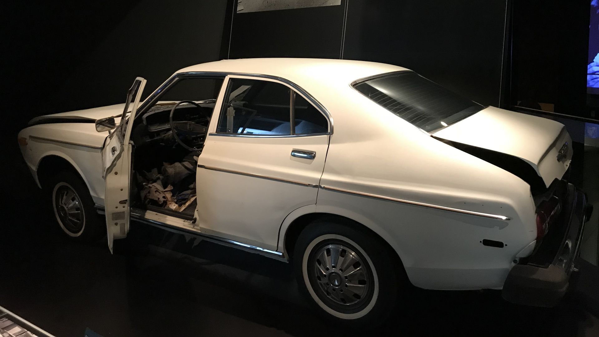 A white 1976 Datsun in a museum display, with its driver side door open, the hood and trunk buckled, and a gaping hole in the driver side floorboard.