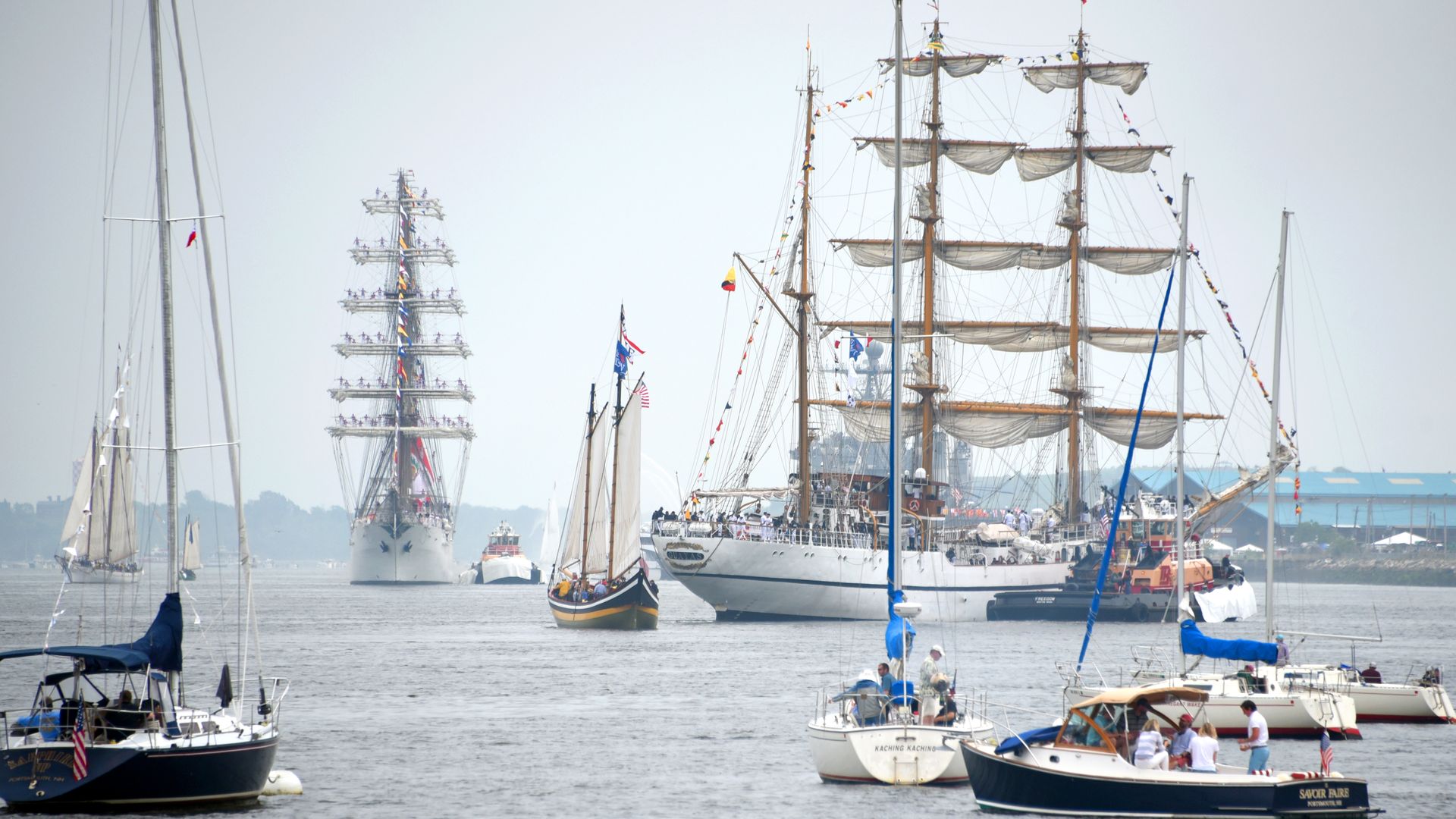 Harbor scene with several tall ships, each having multiple masts and furled sails. Colorful signal flags strung along rigging; smaller boats and crowds visible on the water.