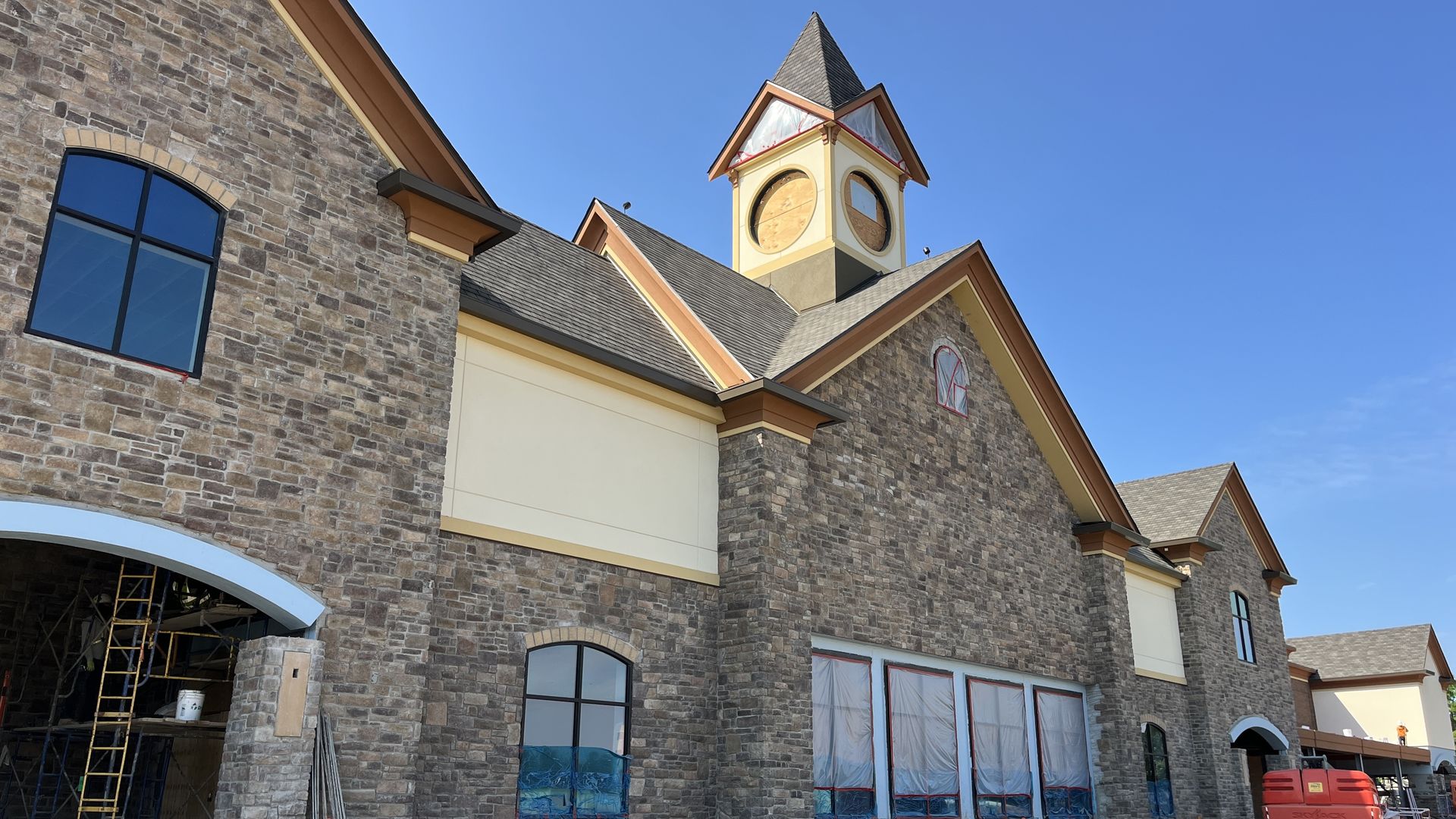 Stone facade building under construction with a yellow clock-tower cupola, arched windows covered in plastic, scaffolding on the left and construction equipment under a clear blue sky.