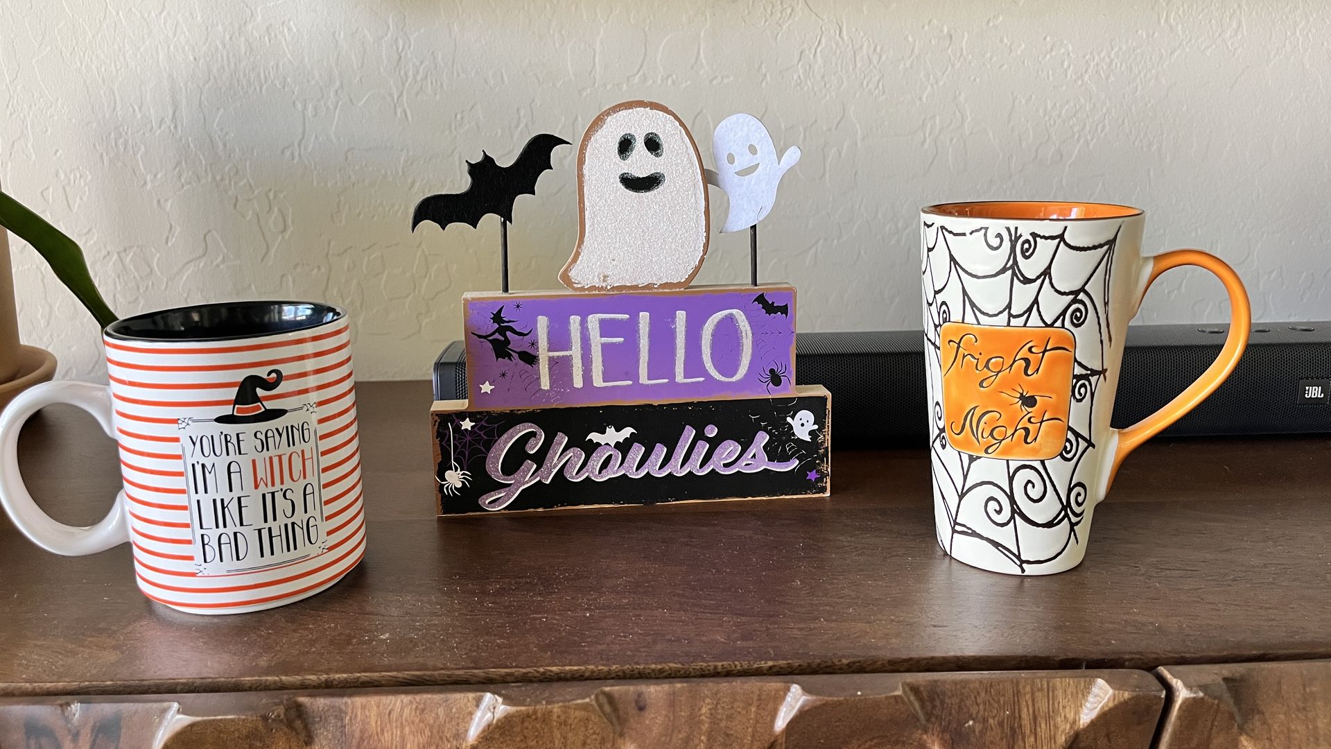 Two Halloween-themed mugs and a decorative wooden block on a wooden surface. Left mug is white with orange stripes and text. Block says 