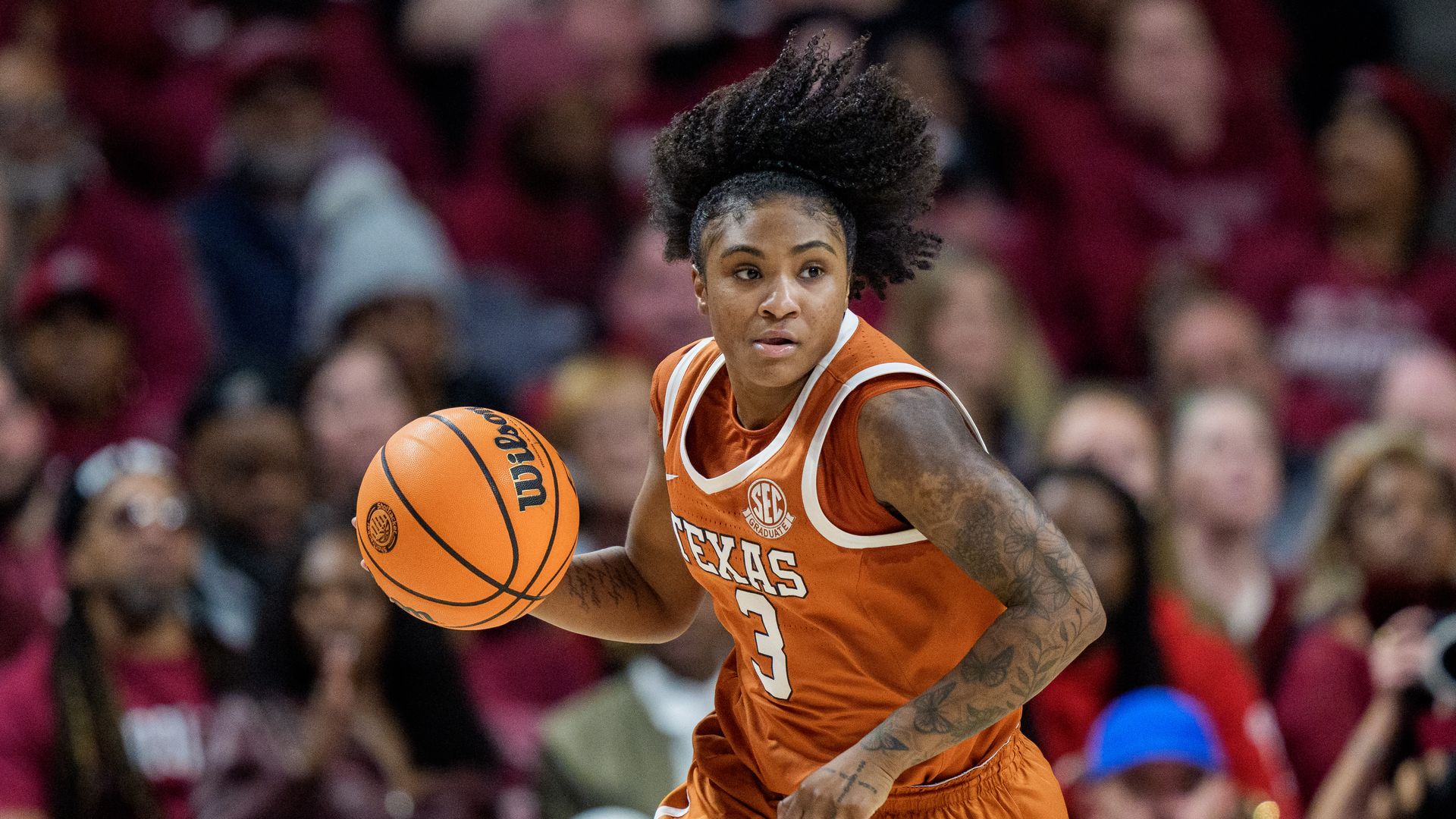 Female basketball player in orange Texas jersey number 3 dribbling a Wilson basketball during a game with a blurred crowd in the background.