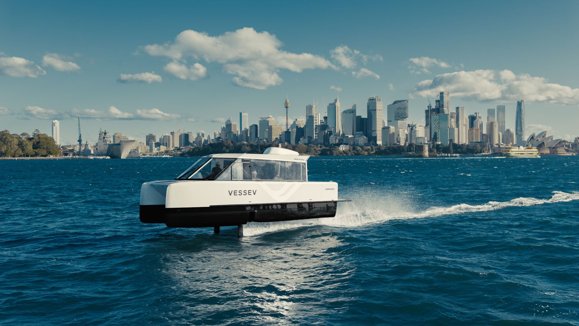 White catamaran labeled "VESSEV" speeds across deep blue water with a distant Sydney skyline, including the Opera House, under a clear blue sky with scattered clouds.