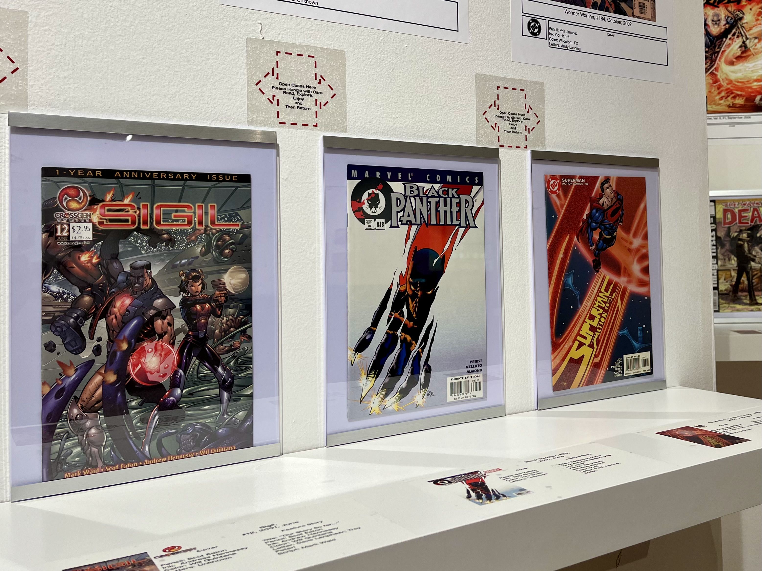 Three comic books on display hanging from a white wall. From left to right: CrossGen's "Sigil," Marvel's "Black Panther" and DC's "Superman"