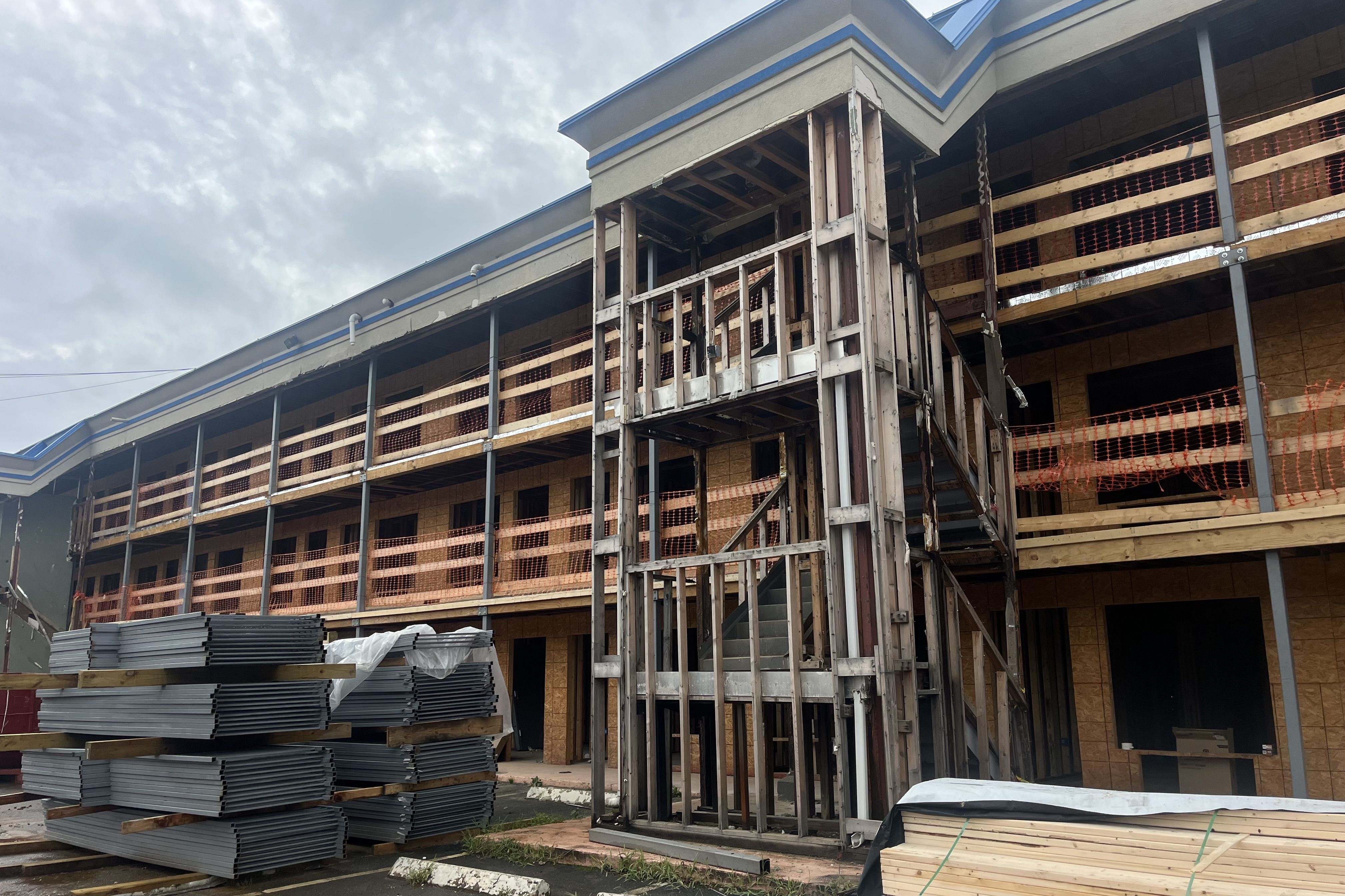 Under construction building with exposed wooden framing and orange safety netting, stacks of metal panels and lumber in front under cloudy sky.