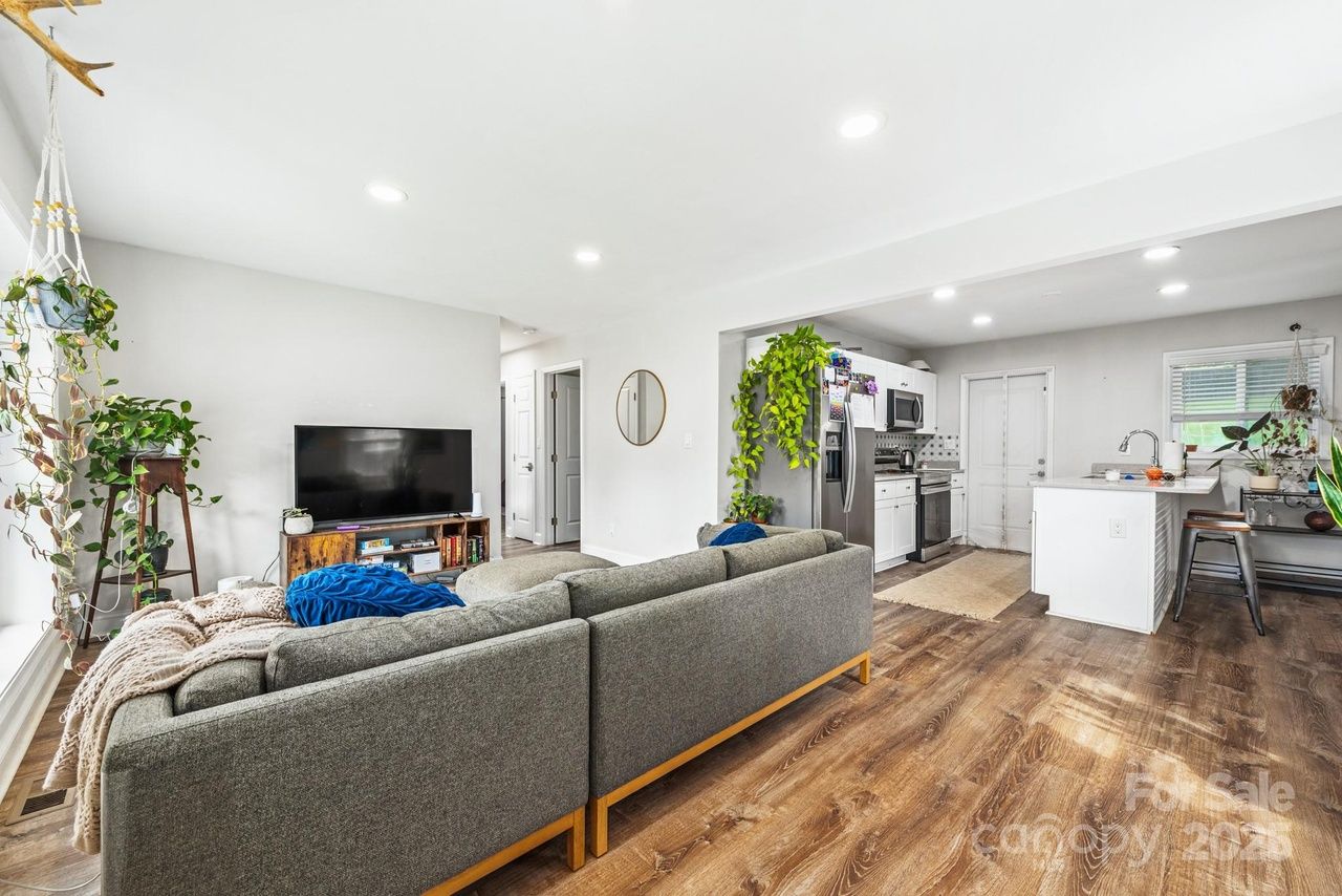 Bright modern living room with gray sectional sofa, wooden floors, large TV on wooden stand, green plants, and an open white kitchen with stainless steel appliances and island.