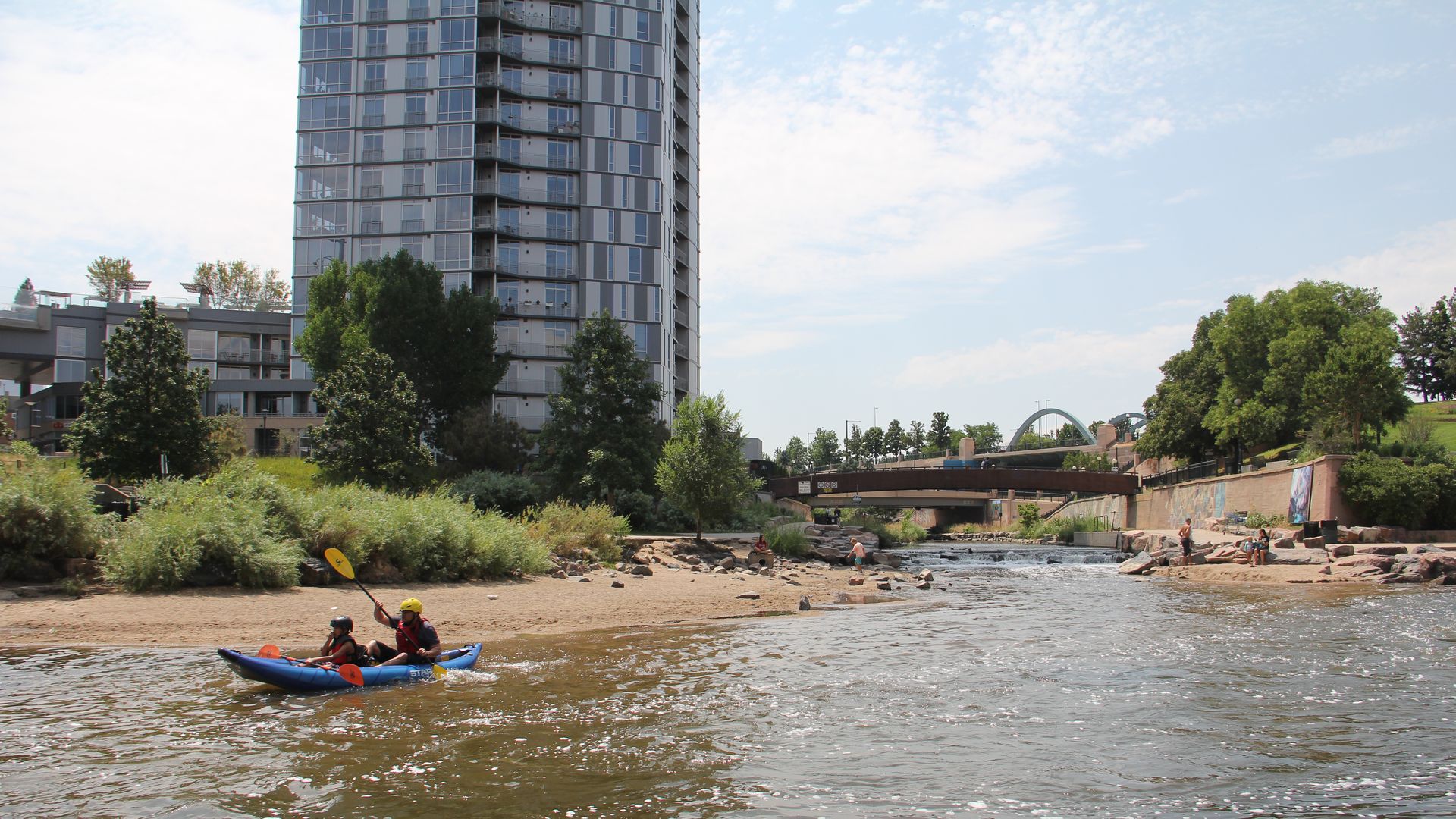 A man and a boy on an inflatable kayak enjoy a riverfront with other people in the background frolicking. 