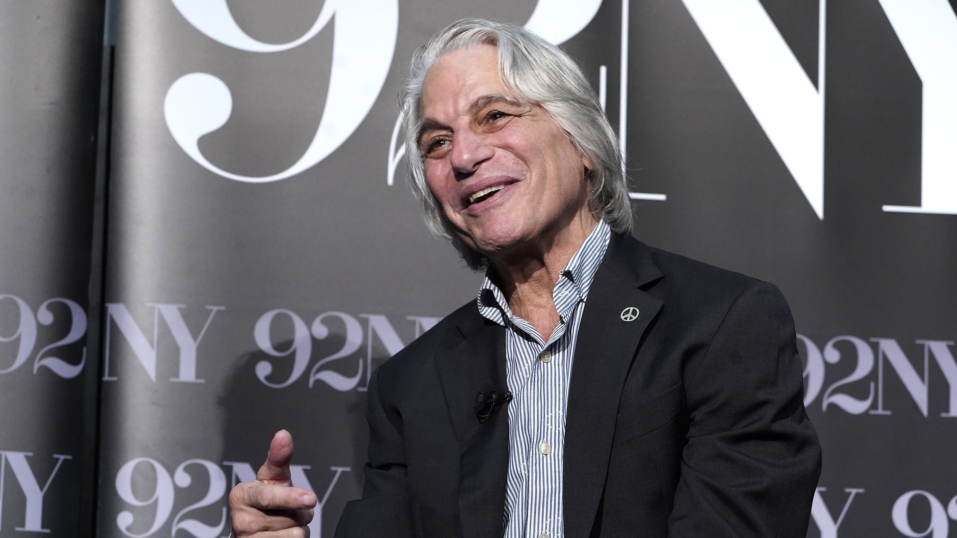Smiling Tony Danza with long gray hair wearing a black blazer and striped shirt, gesturing with hand. 
