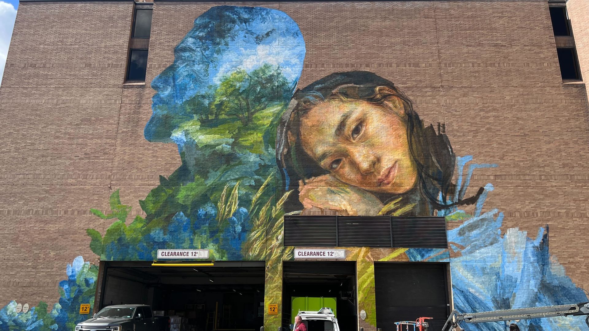 Photo of a mural with a femme figure leaning on an outline of a human filled in with trees and the sky. 