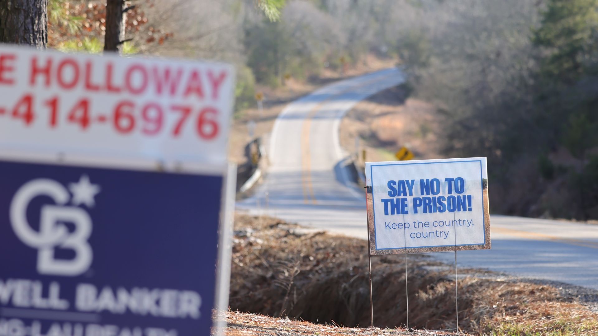 A roadside sign reads "SAY NO TO THE PRISON! Keep the country, country" near a rural road. A real estate sign is visible in the foreground, and a winding two-lane road leads into a wooded area in the background.