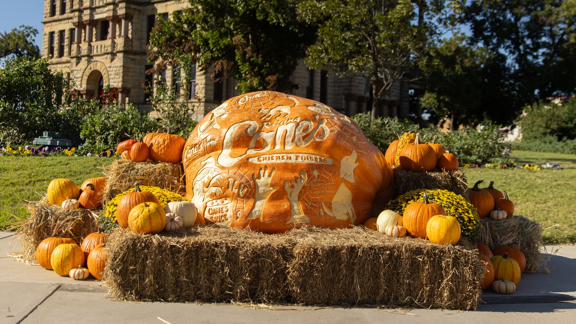 Large carved pumpkin with Raising Cane's Chicken Fingers logo and drawings, surrounded by smaller pumpkins and hay bales, set outdoors near historic stone building under blue sky.