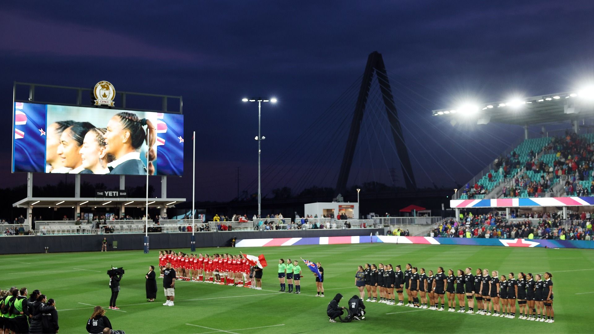 Women rugby players in red and black line up on the grass at CPKC Stadium in Kansas City at night, with a bridge and a large video screen in the background.