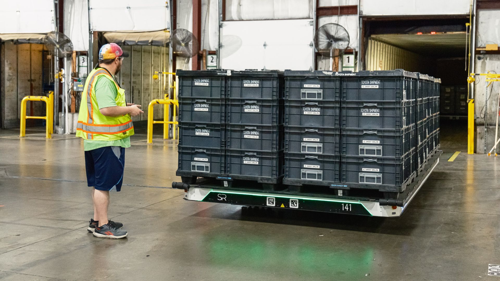 A photo of a barge-like Slip Robotics vehicle carrying crates of goods at a warehouse in Indiana, as a worker in a caution vest looks on.