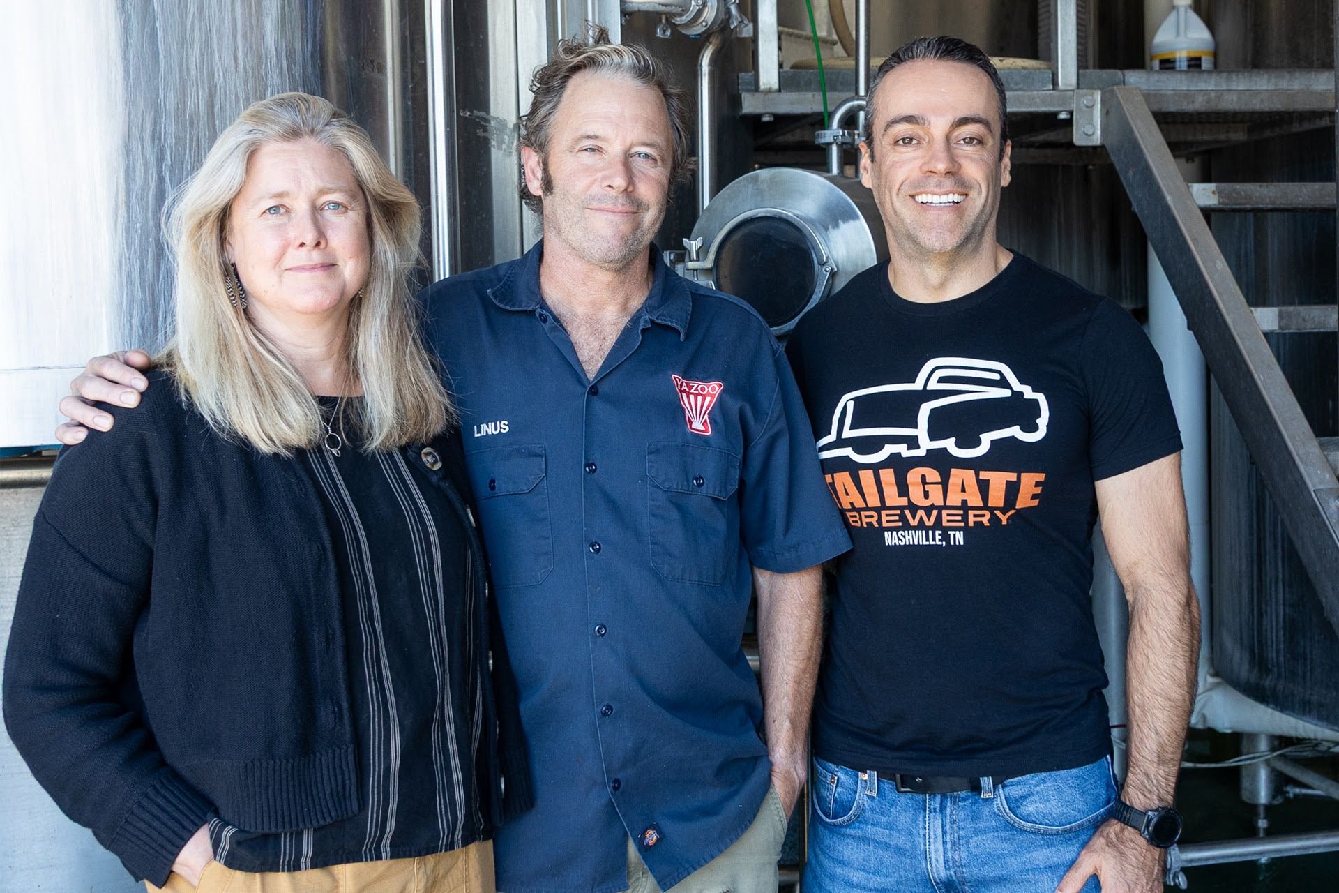 Three people stand in front of industrial brewery equipment: a woman with long blonde hair, a man in a blue work shirt, and a smiling man in a black Tailgate Brewery shirt.