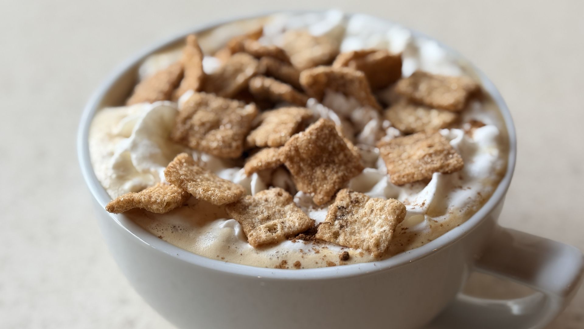Close-up of a white mug with a frothy beverage topped with whipped cream and scattered brown graham-cracker pieces, creating a cozy, dessert-like drink on a light countertop.