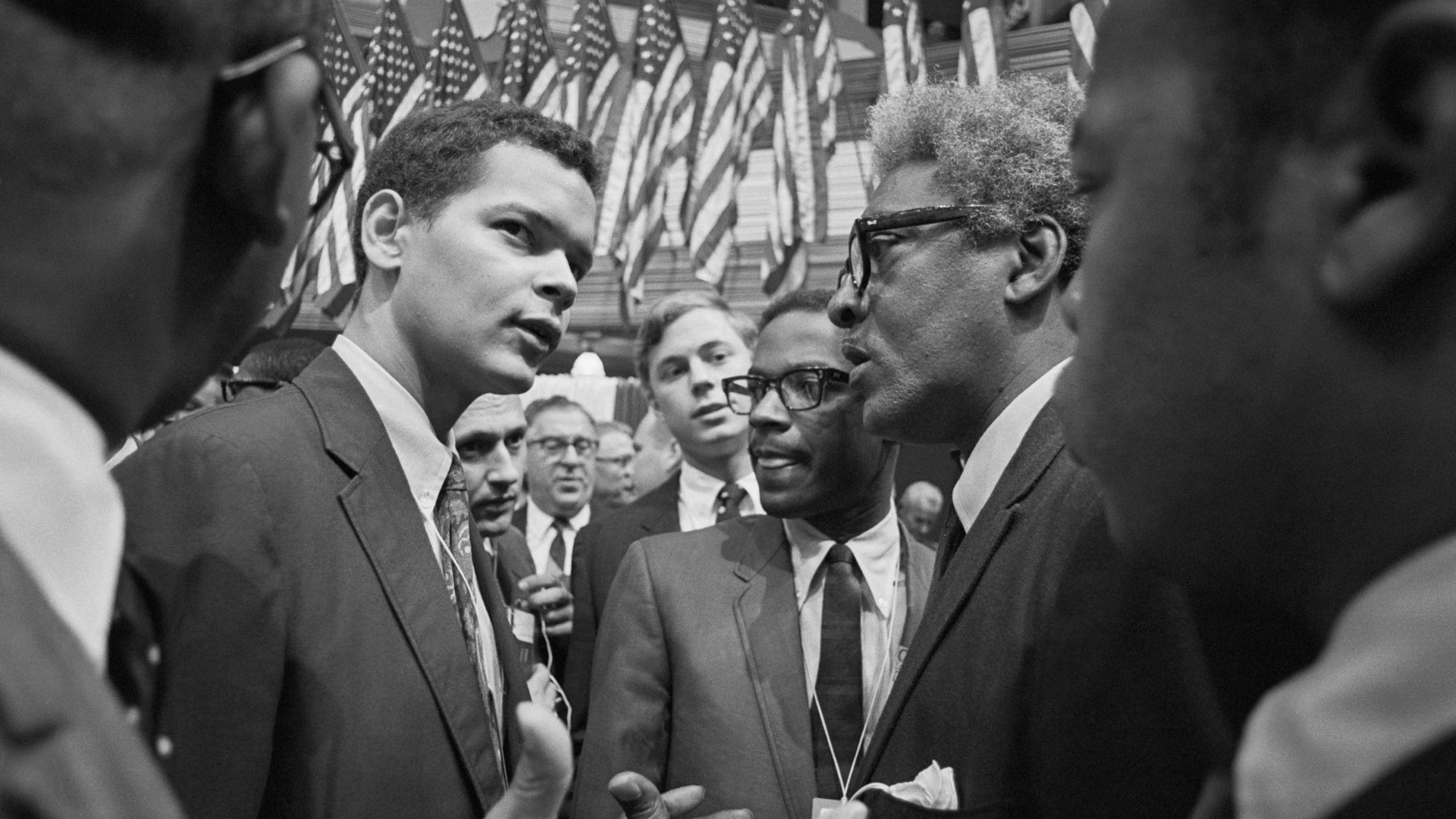 Two individuals wearing suits have a close discussion in a crowded arena with U.S. flags in the background