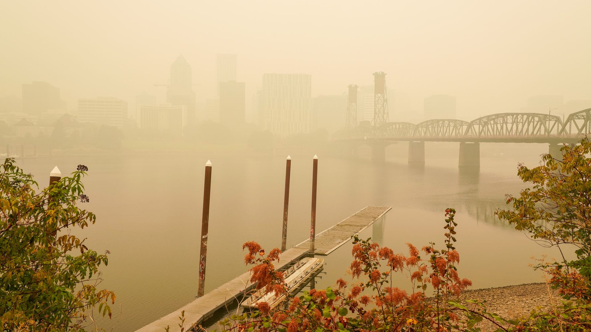 River dock with poles and autumnal plants in foreground, a steel bridge, and hazy city skyline obscured by thick smoke or fog in the background.