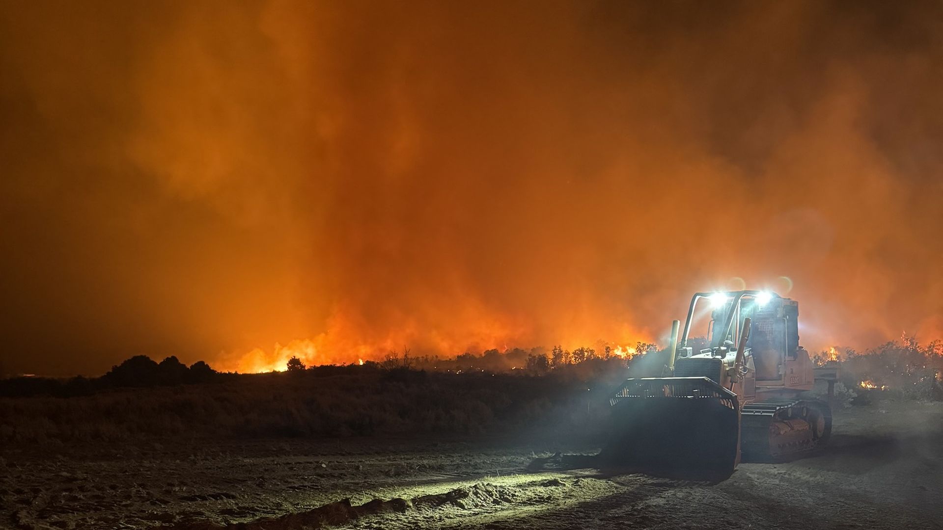 A bulldozer plows over ground near a wildfire at night.