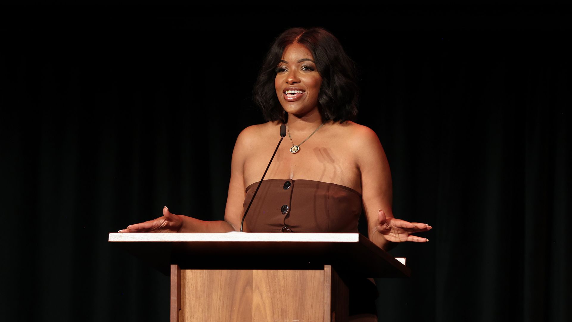 Jasmine Crockett wearing a brown dress and standing at a wooden podium in front of a black backdrop.