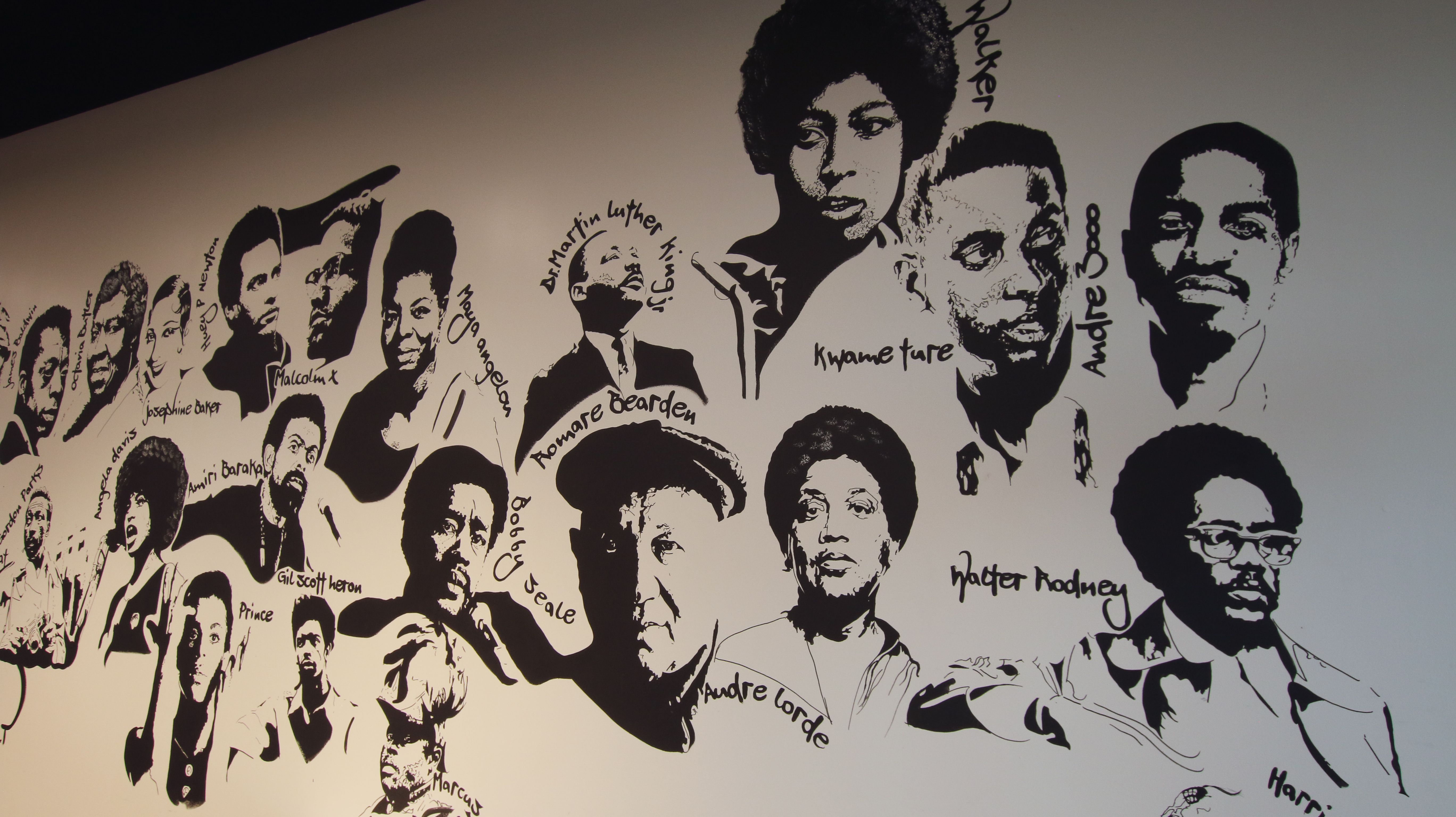 Black-and-white mural on a light wall showing numerous portraits of notable Black figures, with handwritten names around each face (including Malcolm X, Kwame Ture, Walter Rodney, Huey P. Newton).