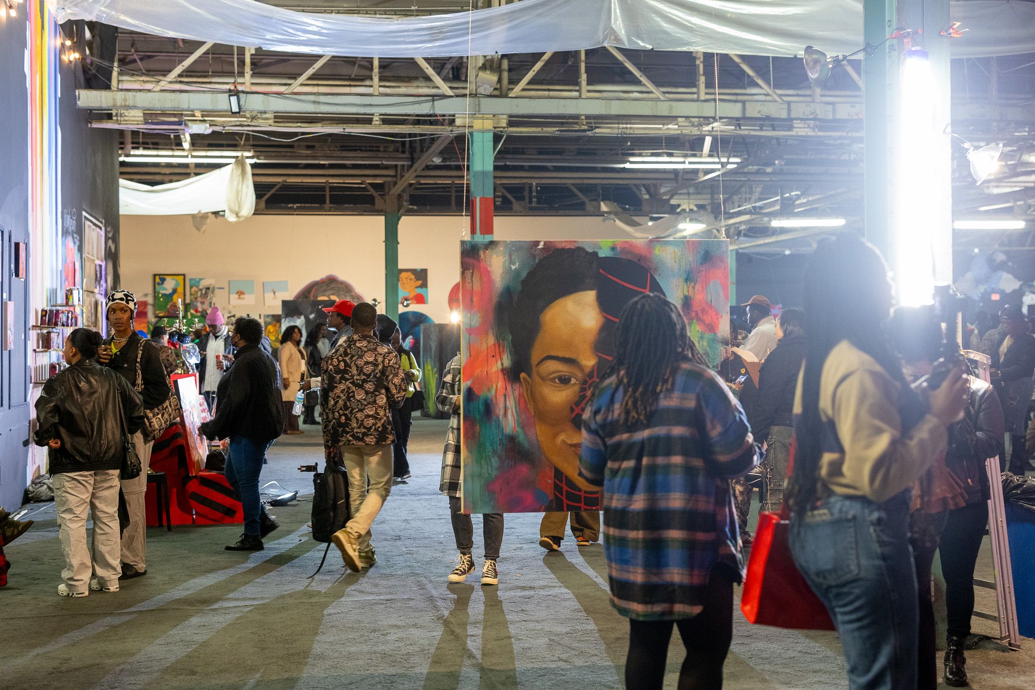 Indoor art event with diverse people viewing colorful paintings and socializing, featuring a large portrait split between a natural face and a red geometric design in center.