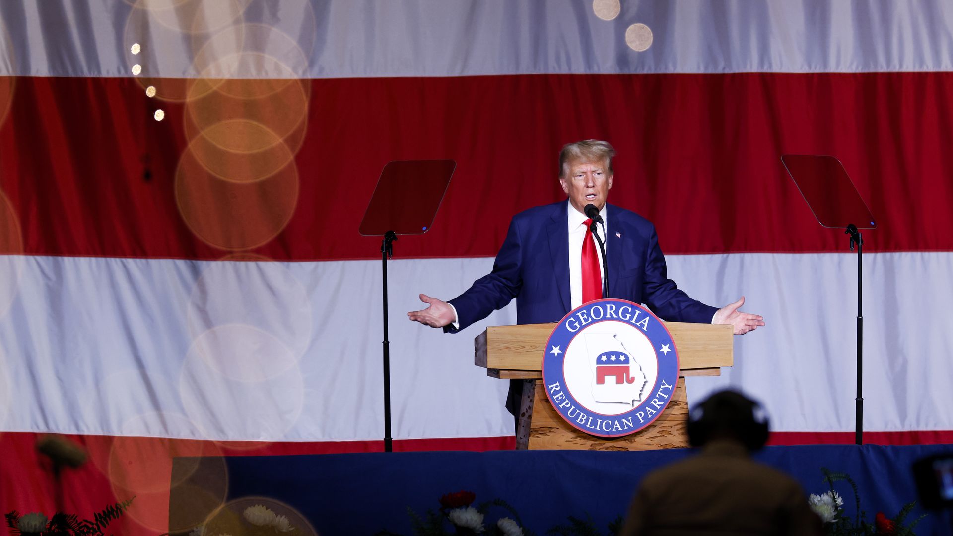 Donald Trump standing at a lectern wearing a blue suit and red tie. His arms are outstretched.