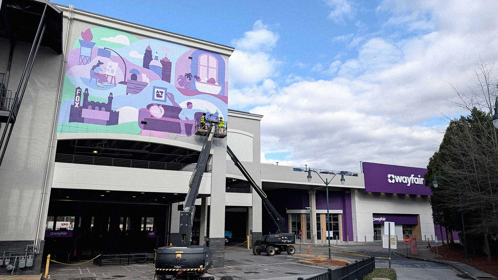 Two workers in a lift painting a colorful mural with buildings and people on a white brick wall of a Wayfair building under a partly cloudy sky.