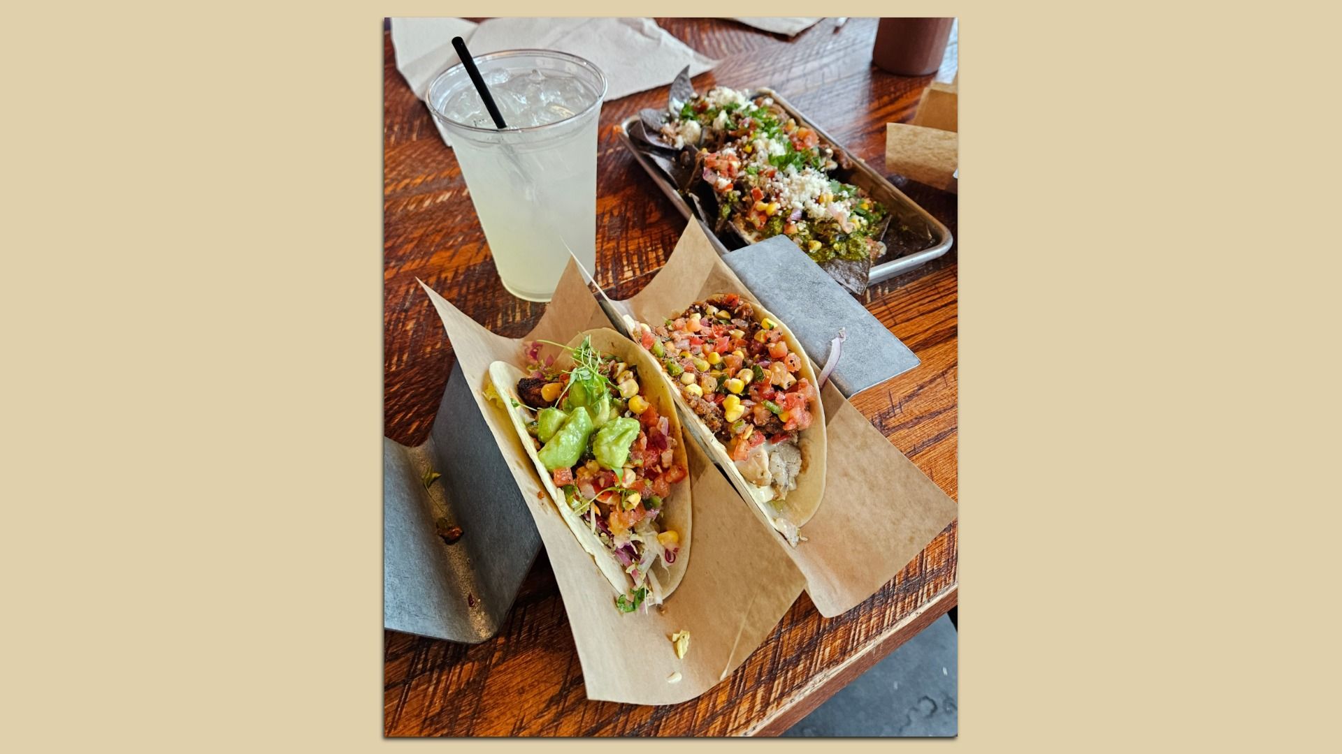Two soft tacos with toppings like guacamole, corn, and salsa on brown paper in a metal holder, a plate of loaded nachos with cheese and vegetables, and a cold drink with a black straw on a wooden table.