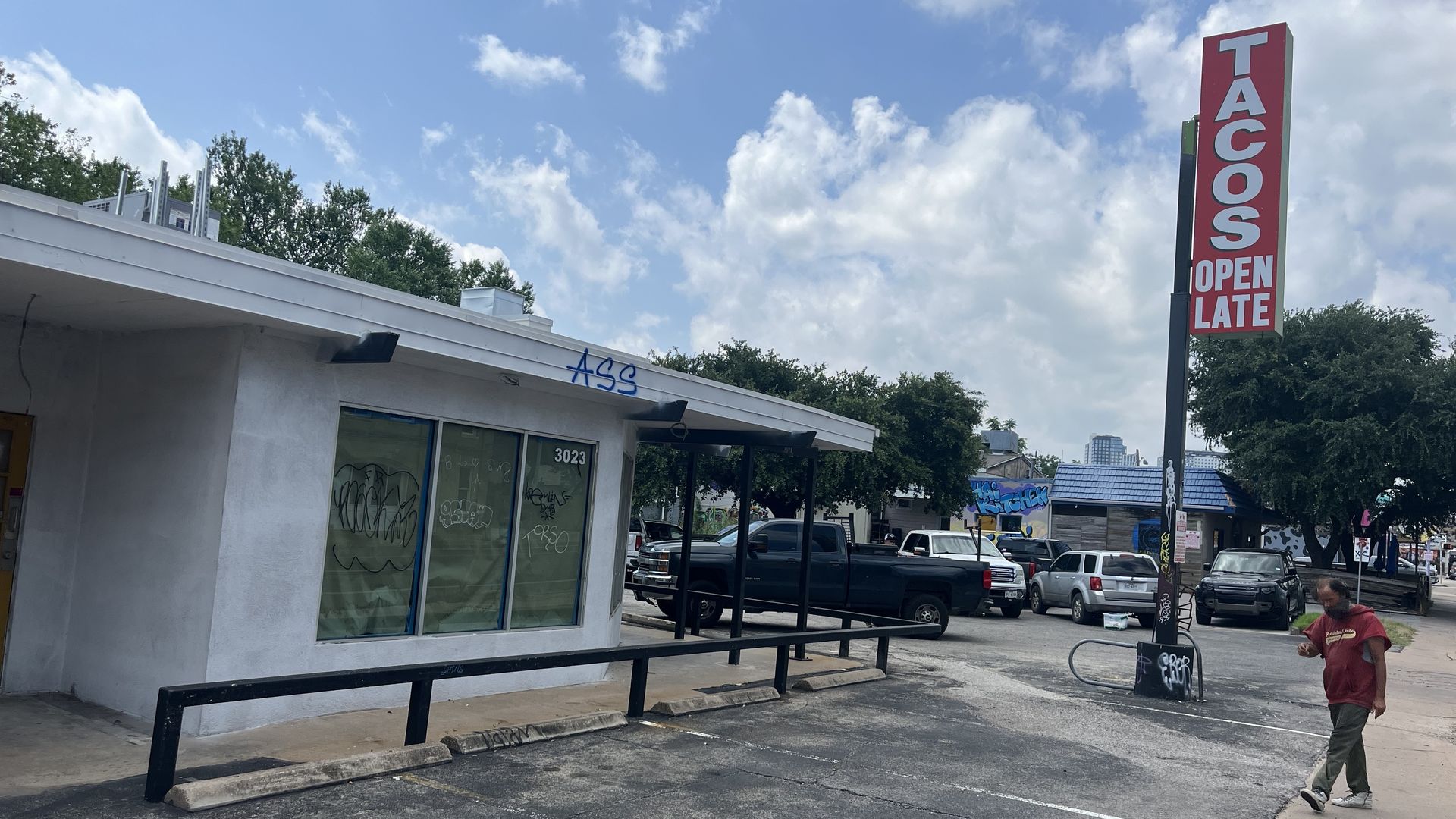 White, single-story storefront with graffiti on the windows. A tall red sign reads "TACOS OPEN LATE" beside a parking lot. Blue sky with clouds; a man in a red shirt walks on the right.