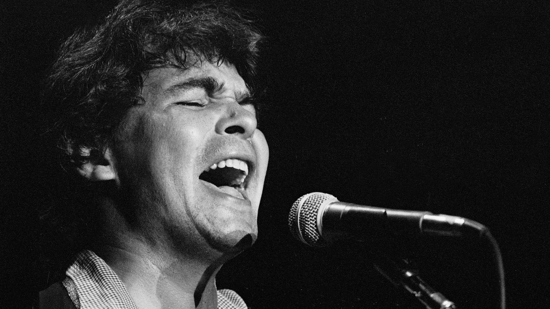 Black-and-white close-up of a male singer passionately singing into a microphone, eyes closed, mouth open, wearing a checkered shirt.