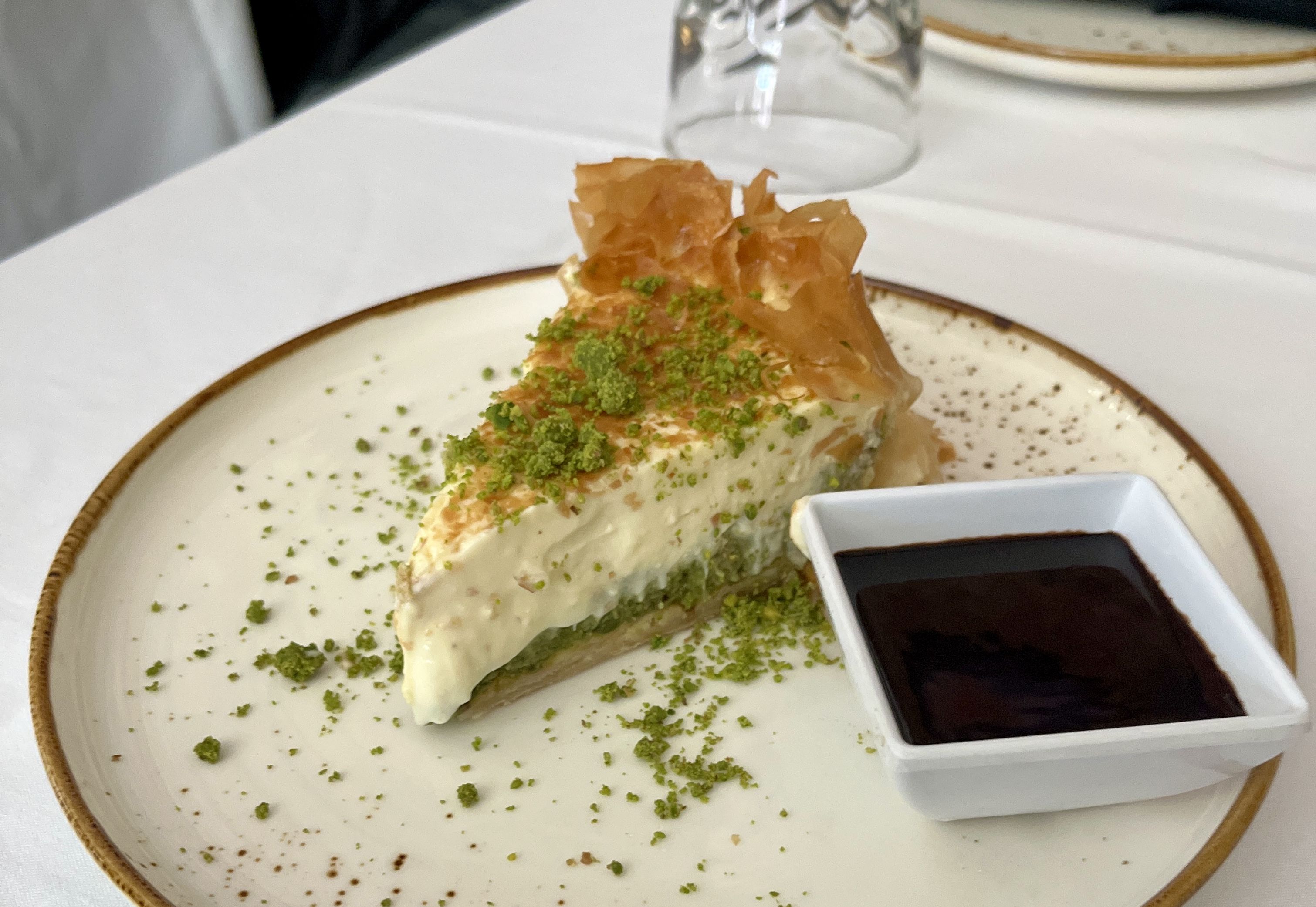 Slice of creamy dessert with green pistachio crumbs on a white plate, served with a small square dish of dark chocolate sauce on a white tablecloth.