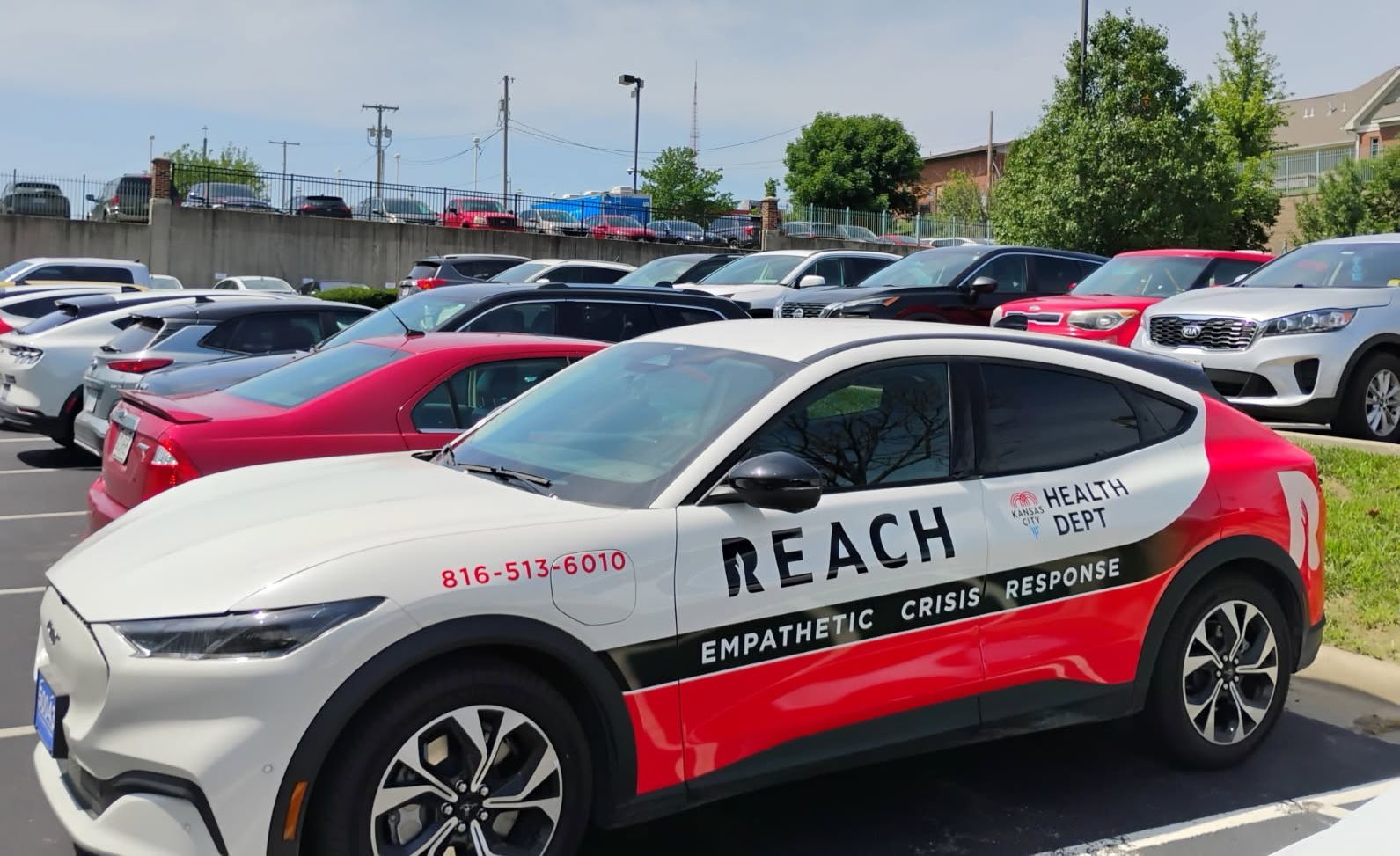 White and red Kansas City Health Dept vehicle with "REACH Empathetic Crisis Response" text parked in a lot with other cars and buildings in the background.